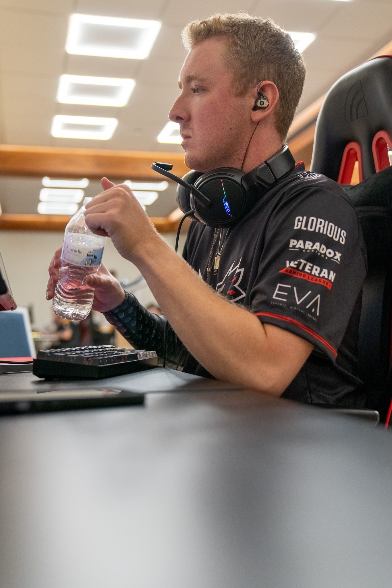 Esports player wearing headphones and gaming jersey holds water bottle while seated at desk with computer.