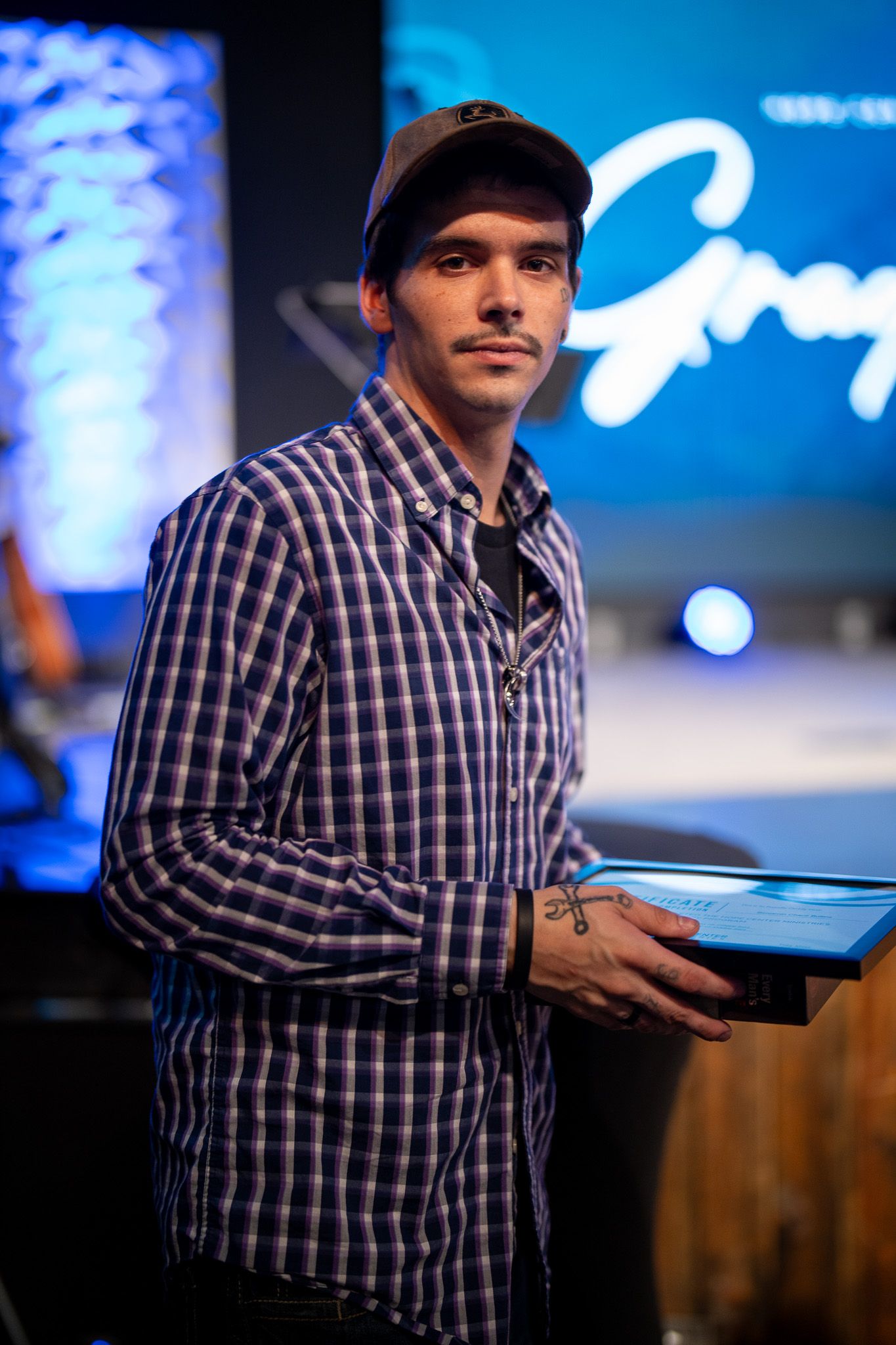 Server holding tablet device in plaid shirt at restaurant counter with blue illuminated signage in background.