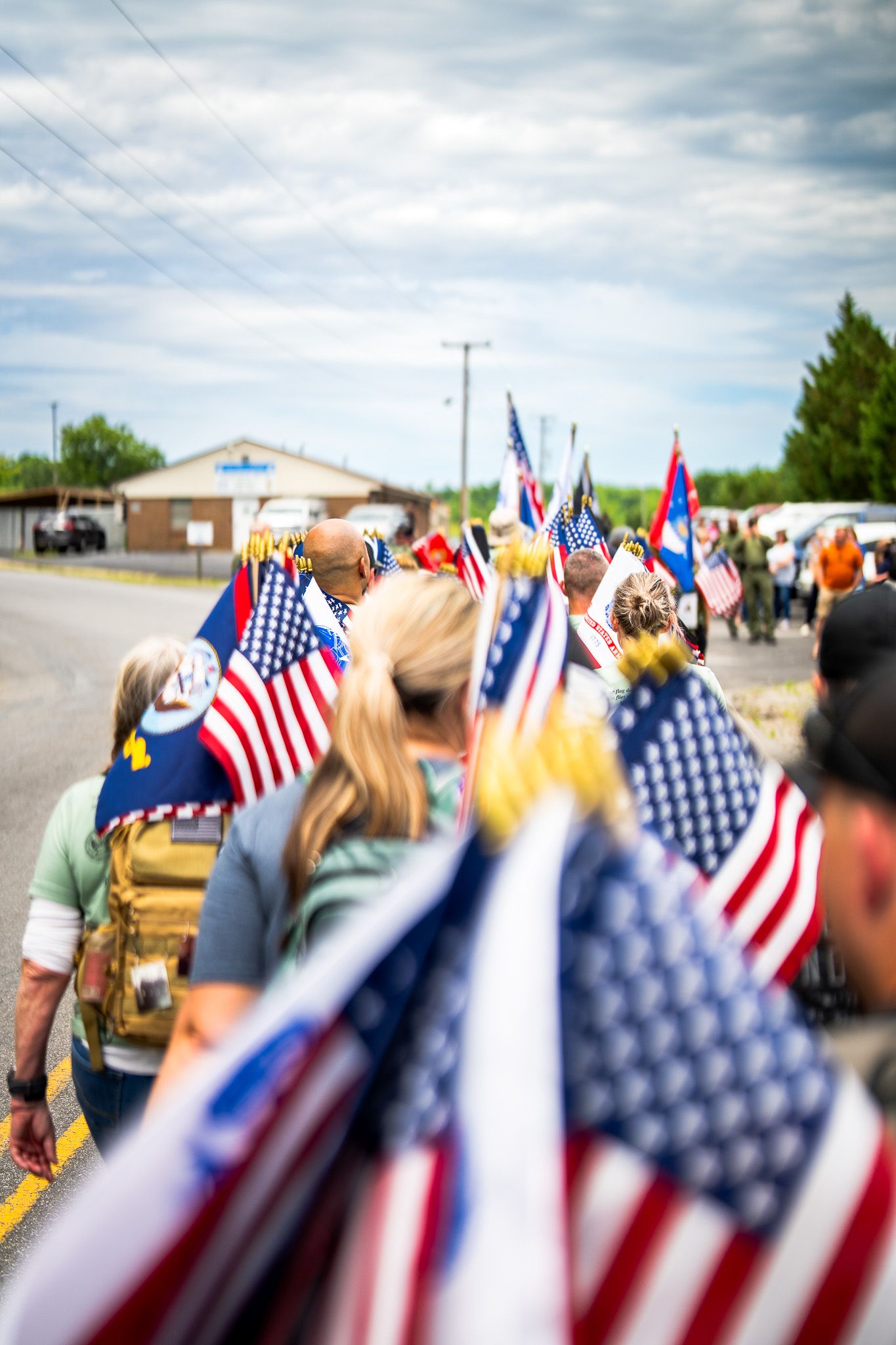 Crowd gathered at patriotic event with multiple American flags waving in foreground against cloudy sky.
