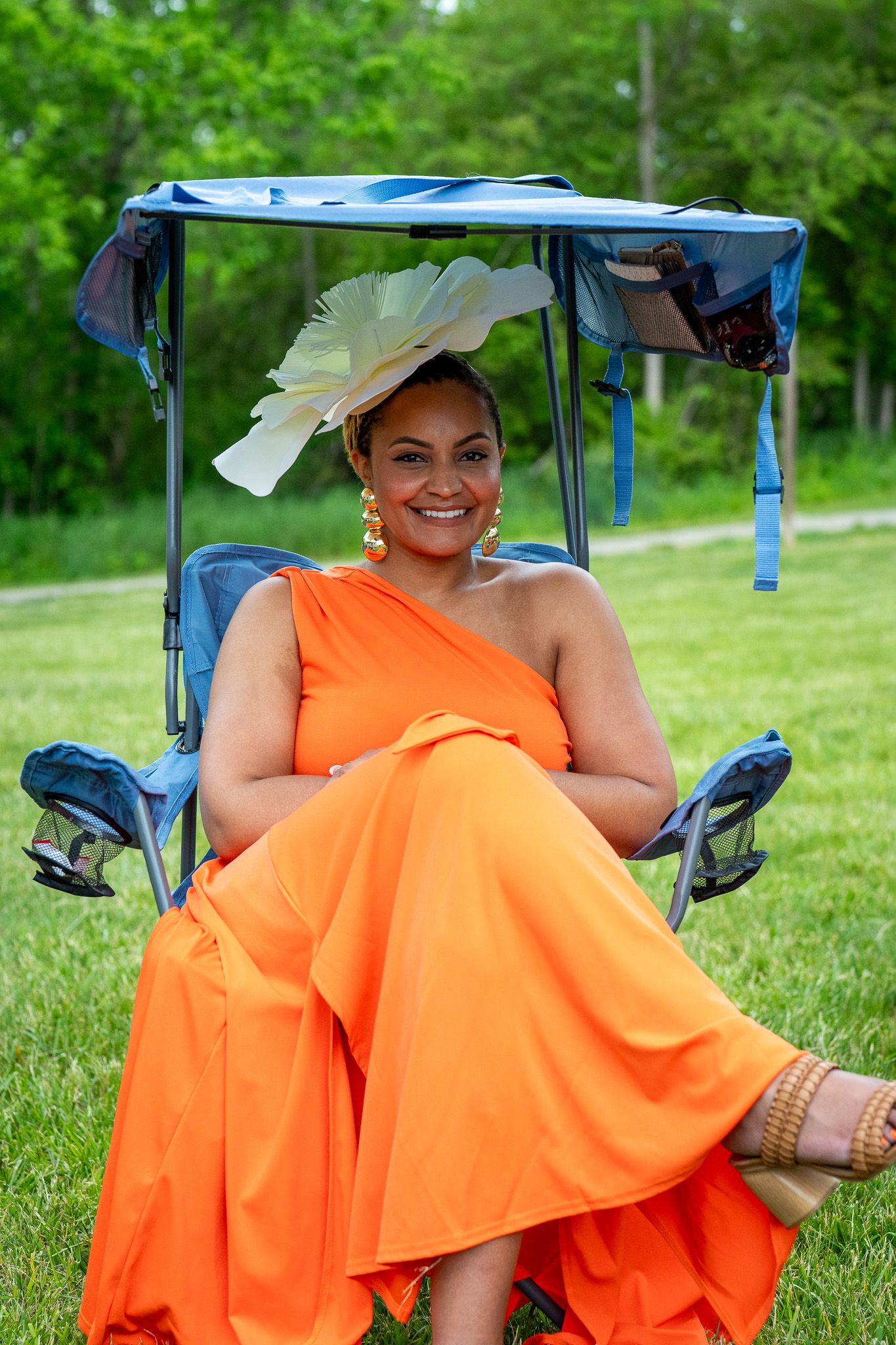 Someone in an elegant orange one-shoulder dress and statement hat sits smiling in a shaded outdoor chair on grass.