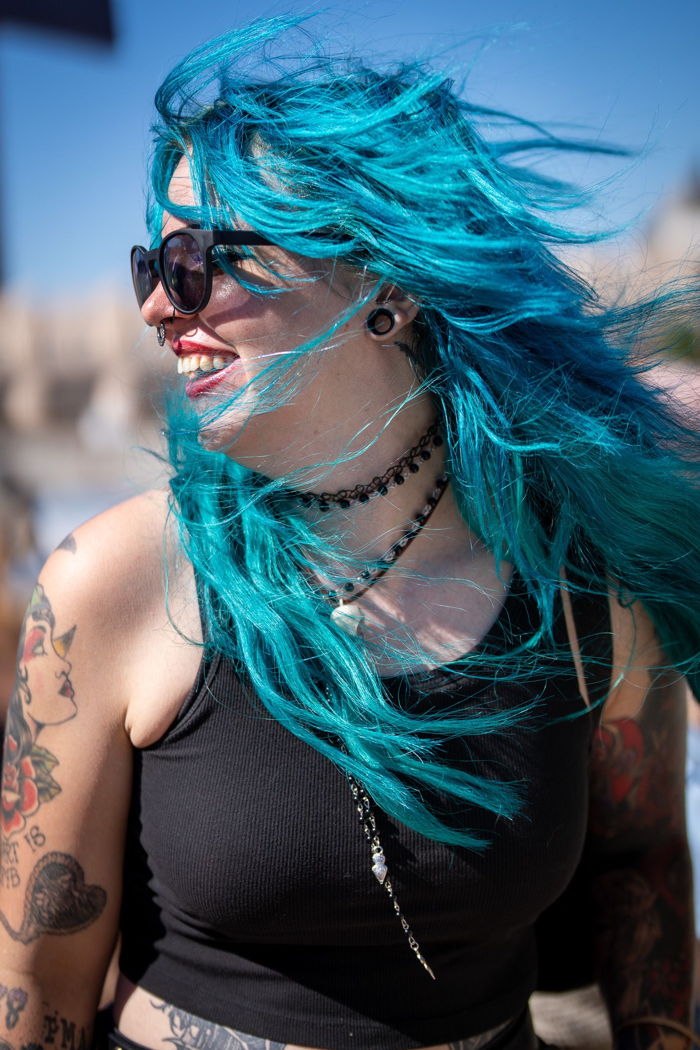 Person with vibrant turquoise hair wearing sunglasses showcases tattoos in black tank top against blurred background.