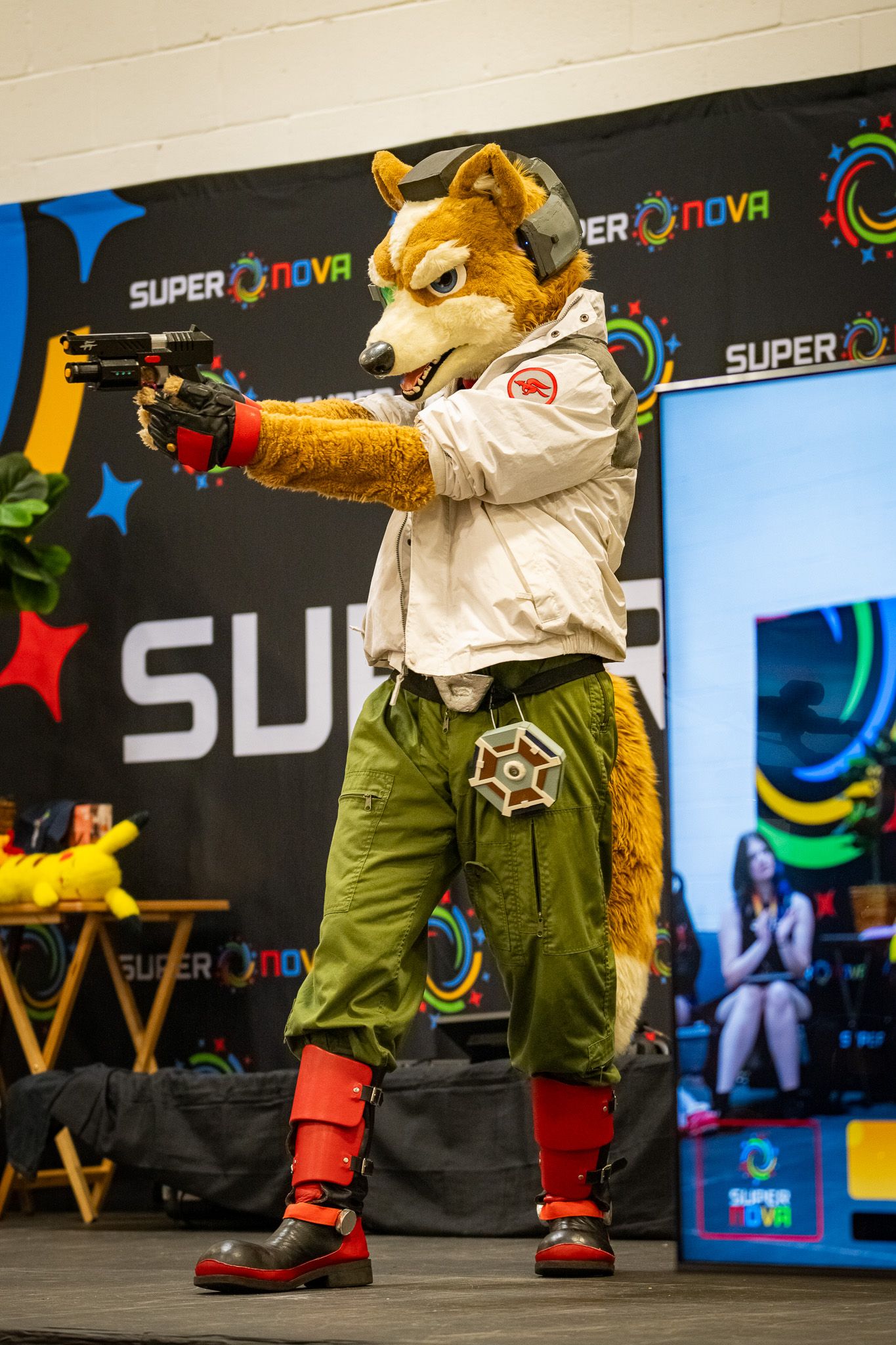 A fox fursuit character in military-style attire aims a prop gun at a gaming convention.