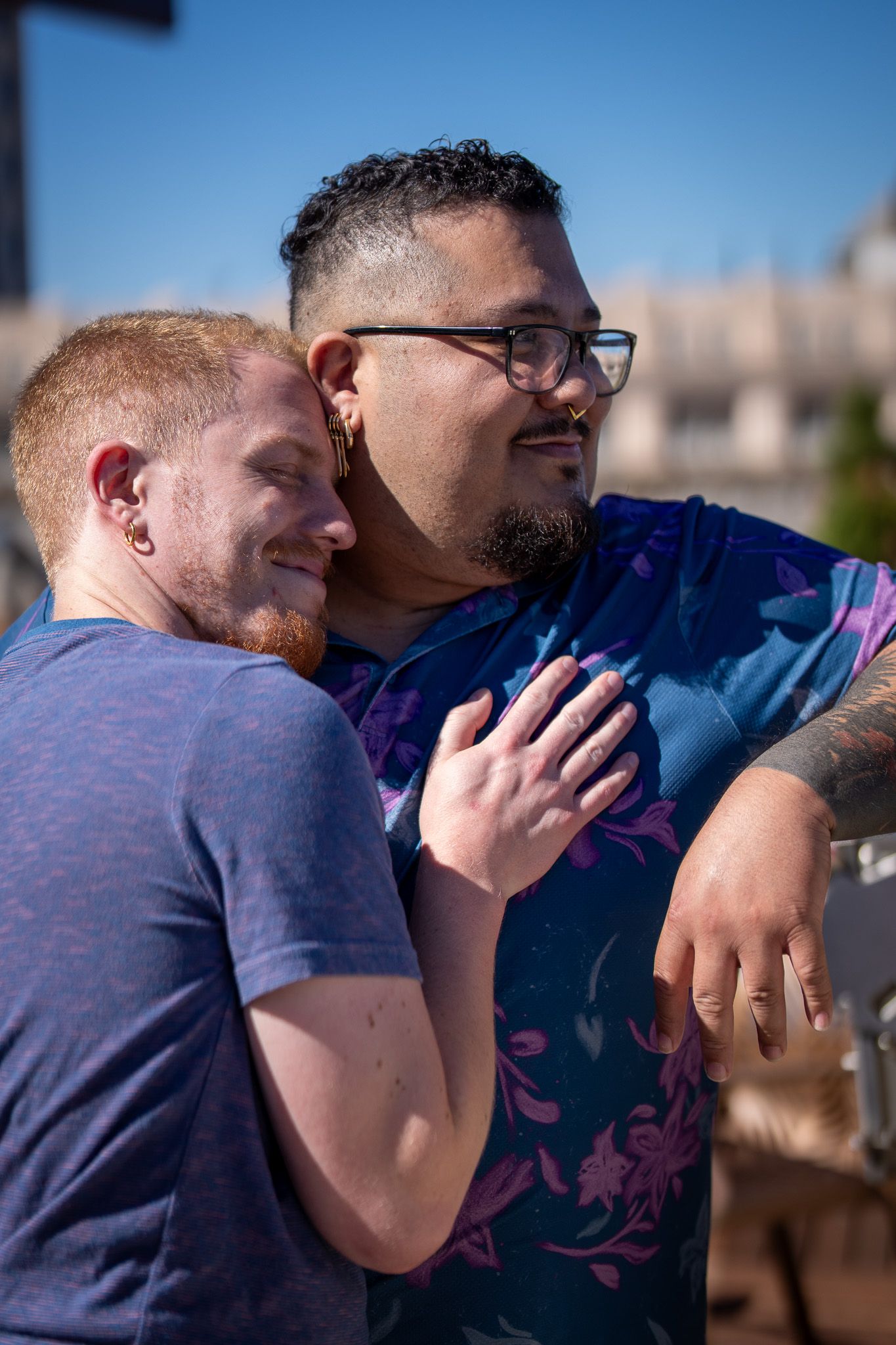 A couple embraces outdoors on a sunny day, one wearing glasses and a floral shirt while the other wears a purple t-shirt.