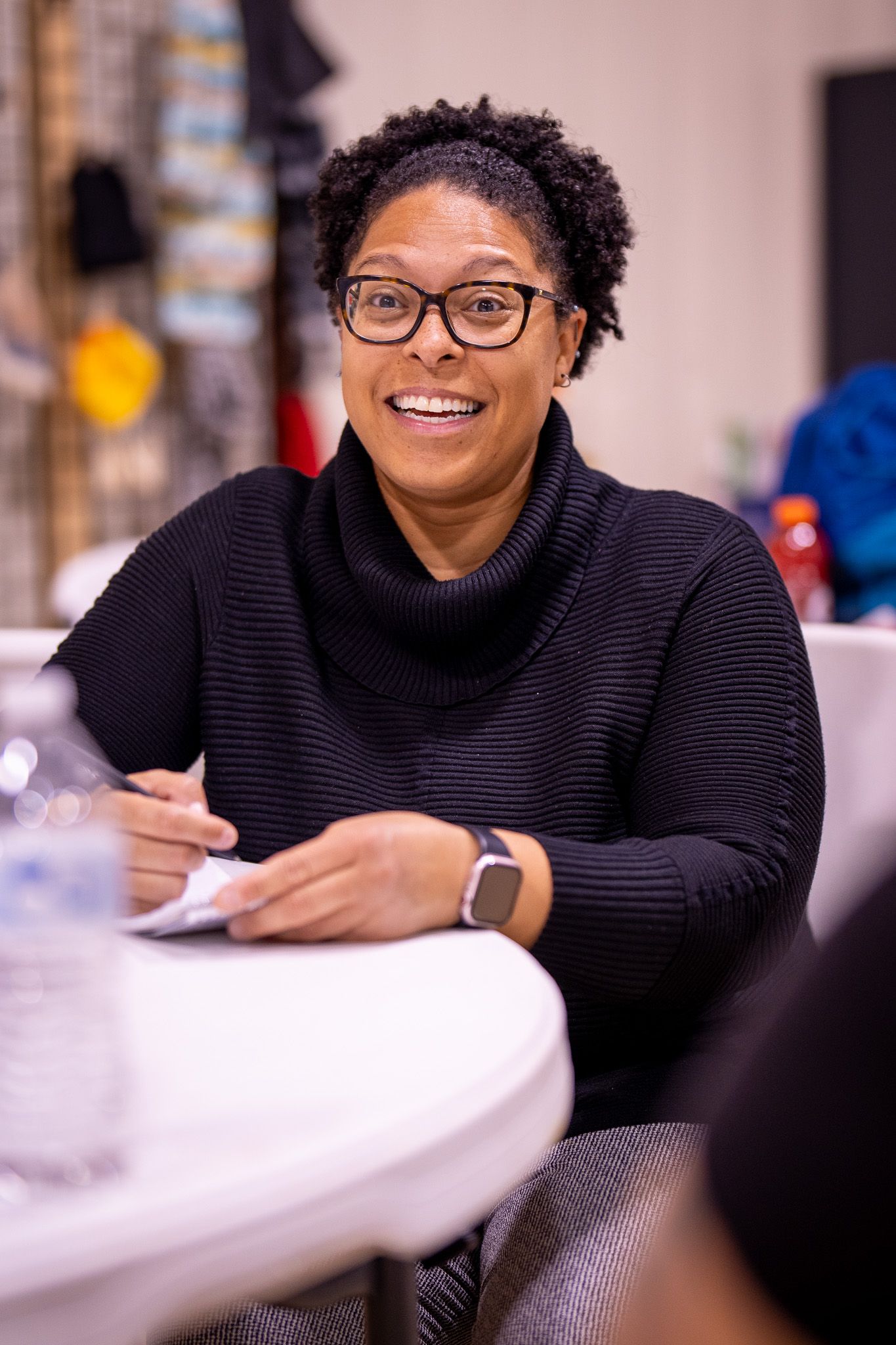 Professional wearing glasses and black turtleneck sweater sits at table with smart watch, smiling warmly during meeting.