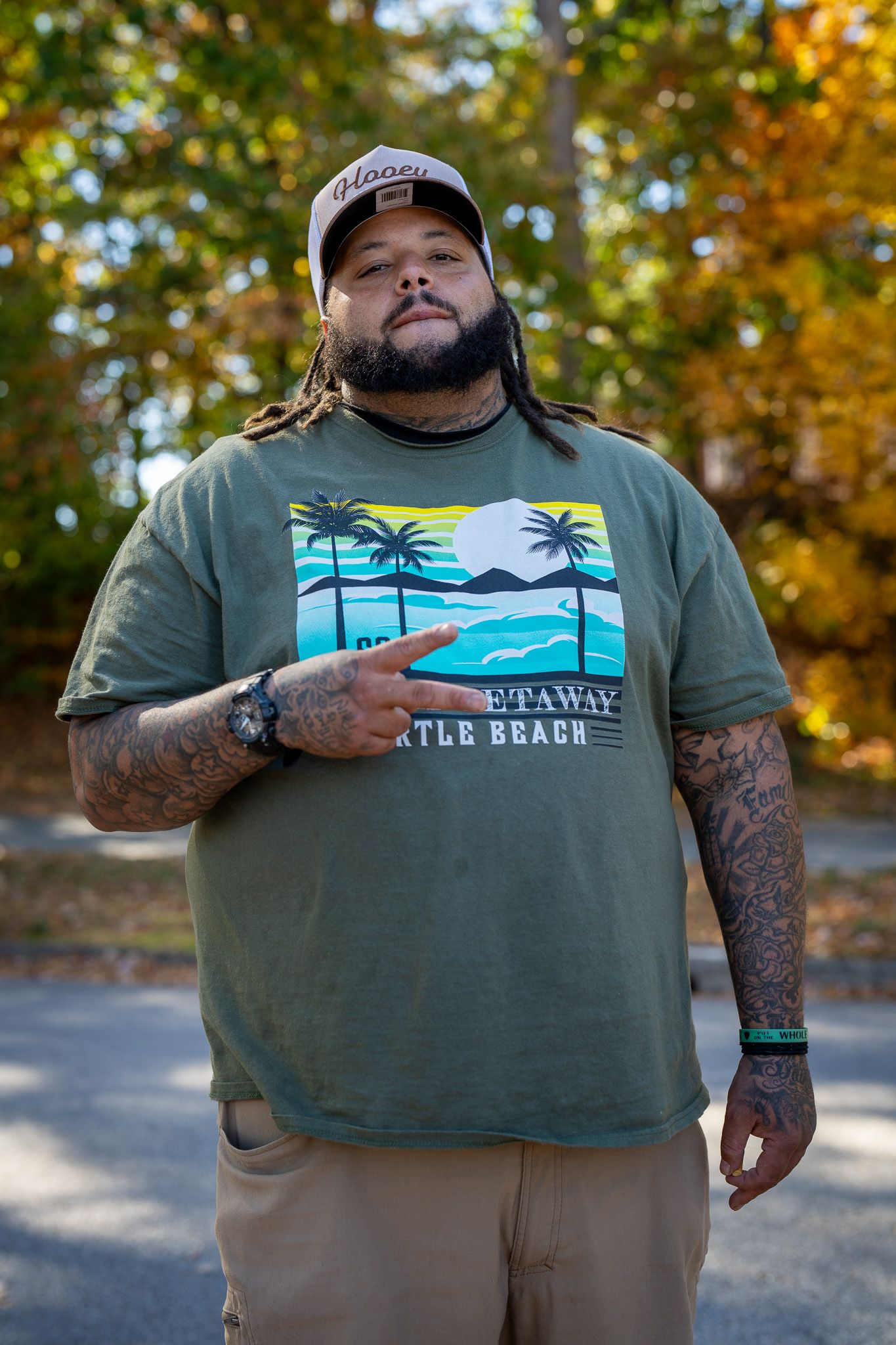 Person wearing a green graphic t-shirt with beach scene and khaki shorts poses outdoors among fall foliage.