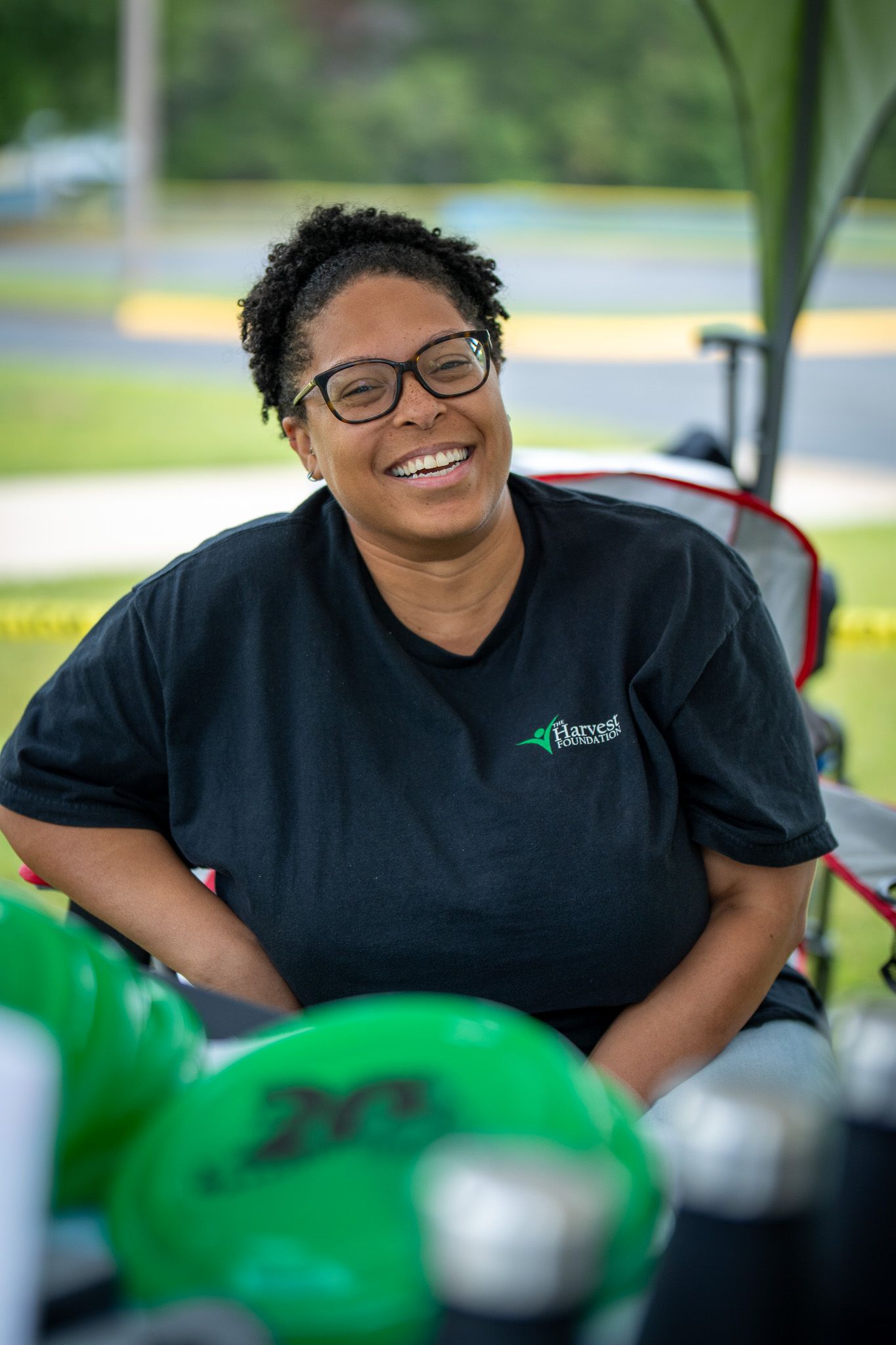 Person in black polo shirt smiling warmly while working outdoors with green equipment visible in foreground.