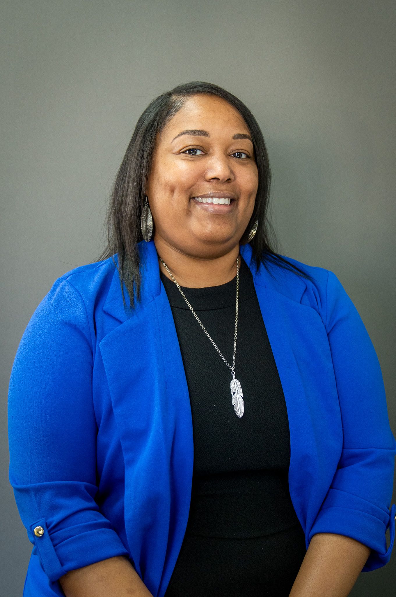 Professional portrait of someone wearing a bright blue blazer and feather necklace against a gray background.