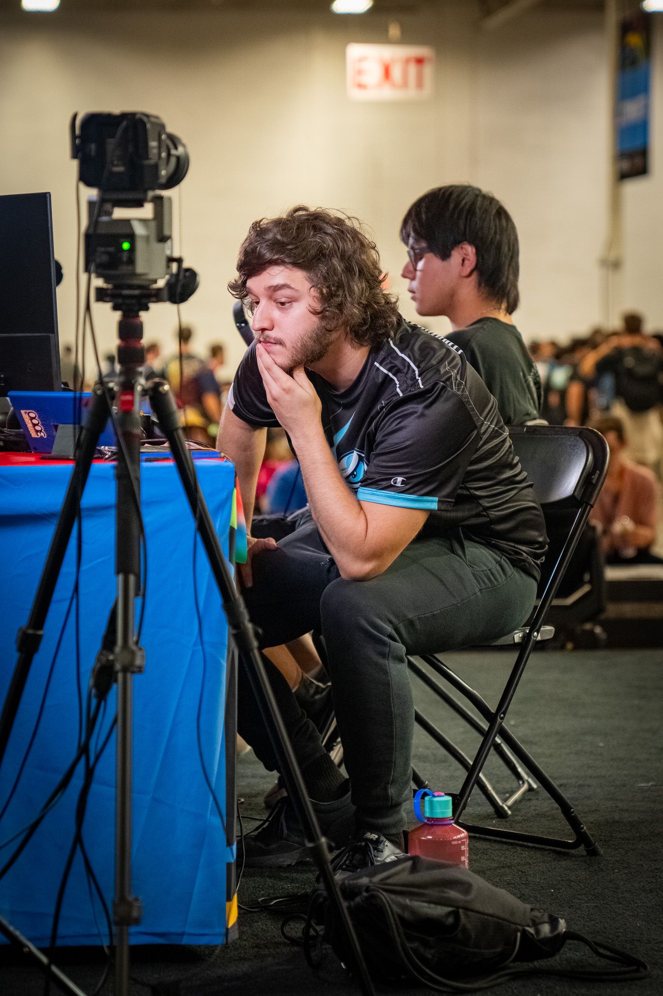 People using cameras and equipment at a gaming or esports event with a blue table and folding chairs in frame.