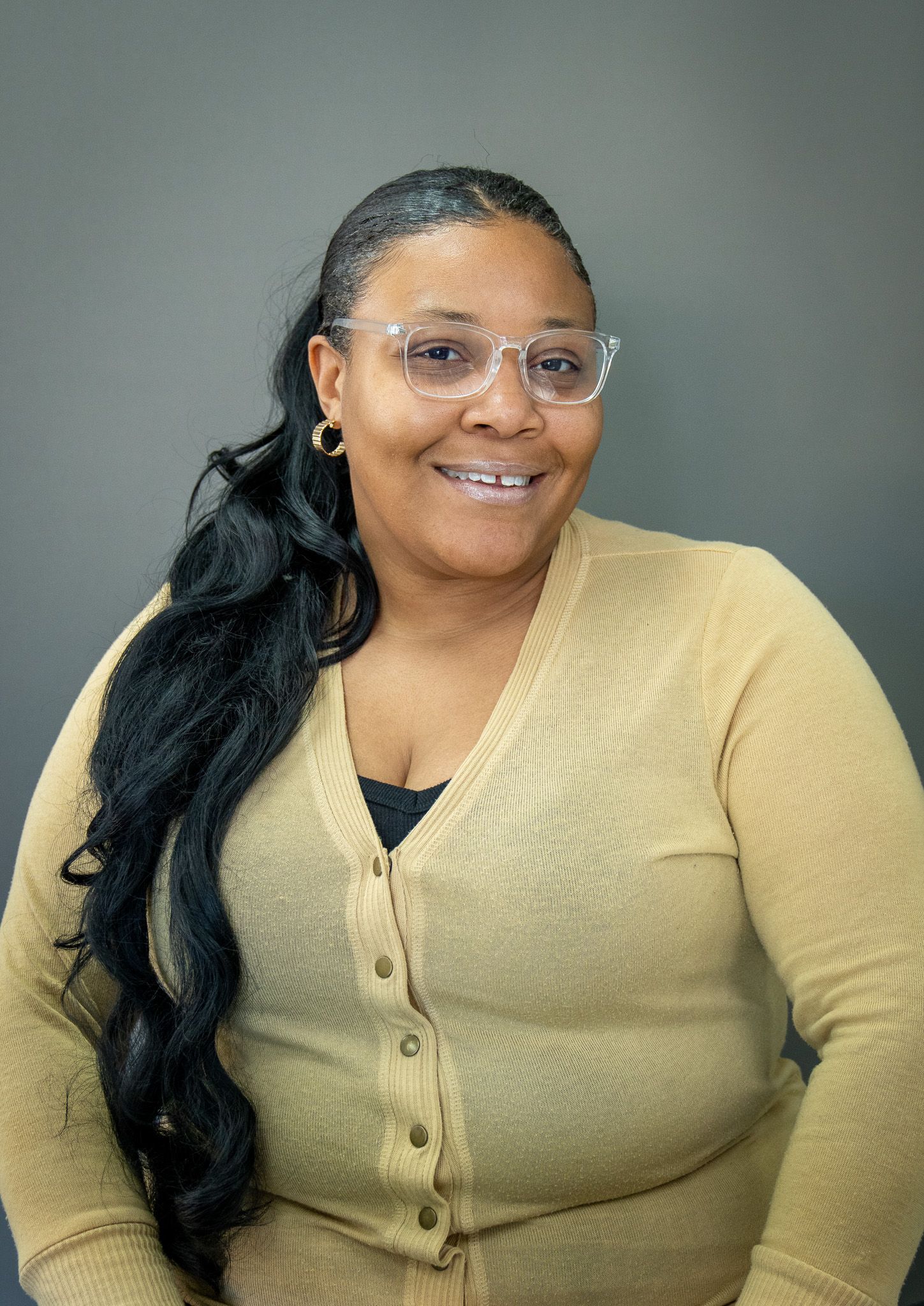 Professional headshot of someone in a tan cardigan and glasses smiling warmly against a gray background.