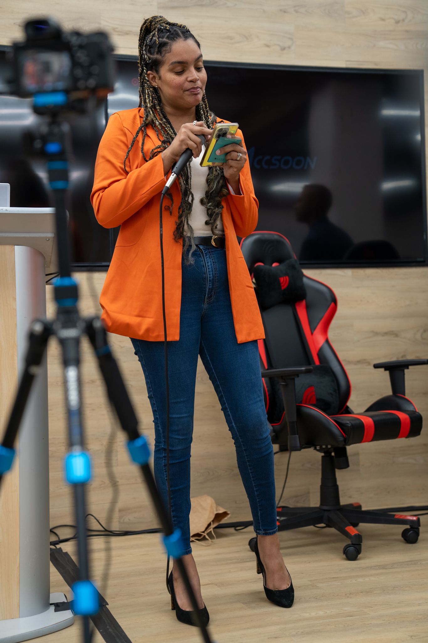 A streamer in an orange cardigan and jeans checking their phone while standing in a gaming setup with camera equipment nearby.