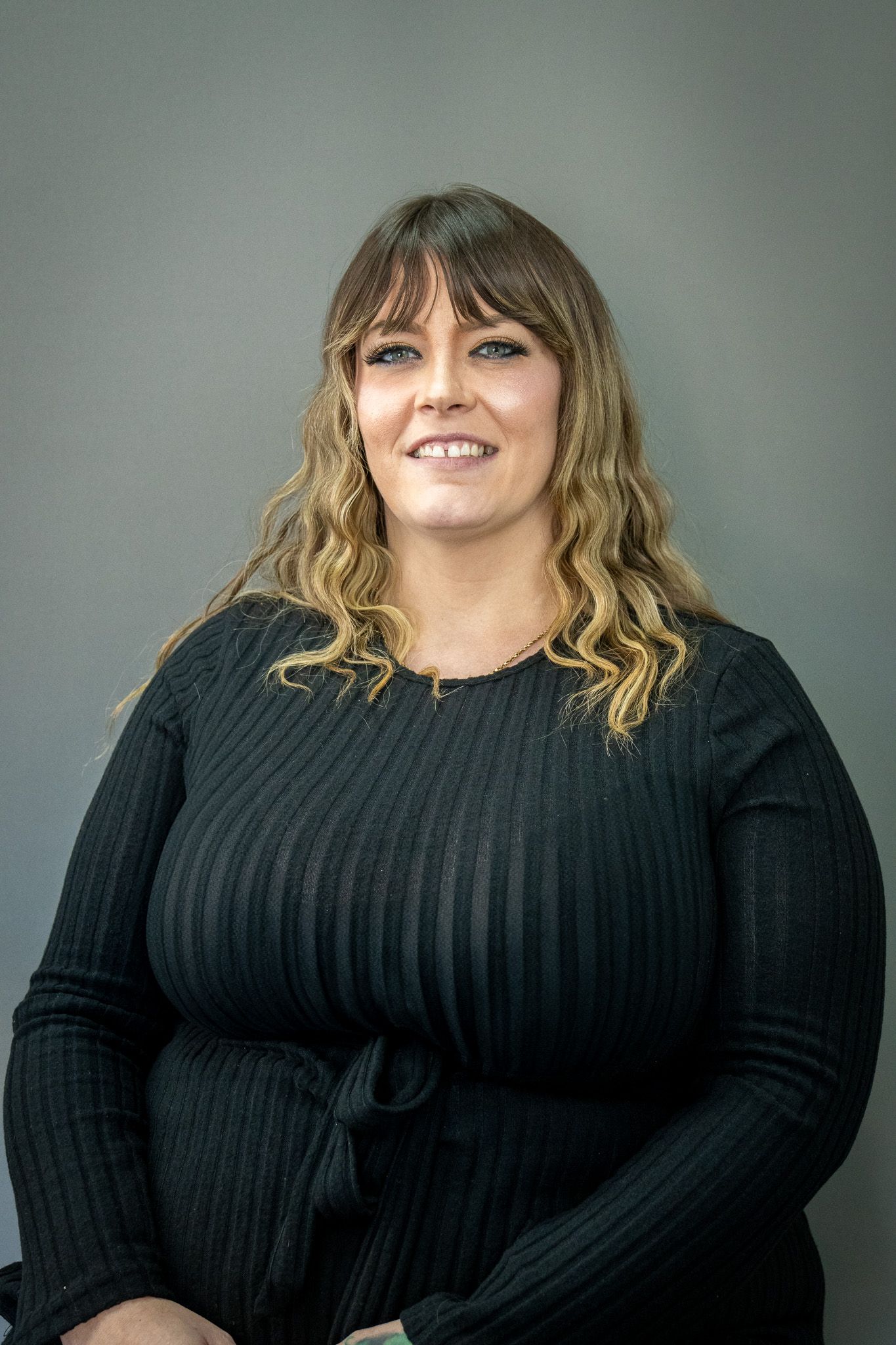 Professional portrait of person in black ribbed sweater with long wavy hair against grey background.