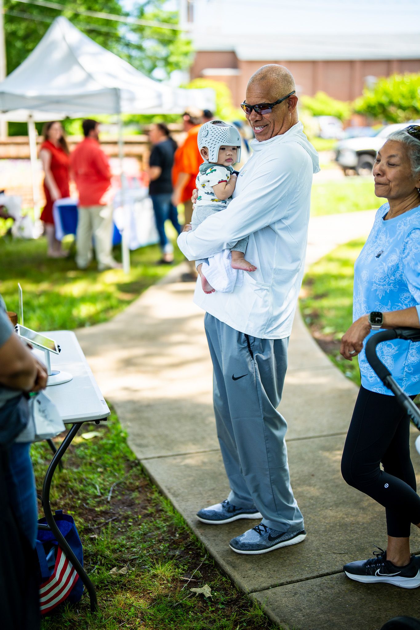 People gathered at an outdoor community event on a sunny day with tents and refreshments visible in the background.