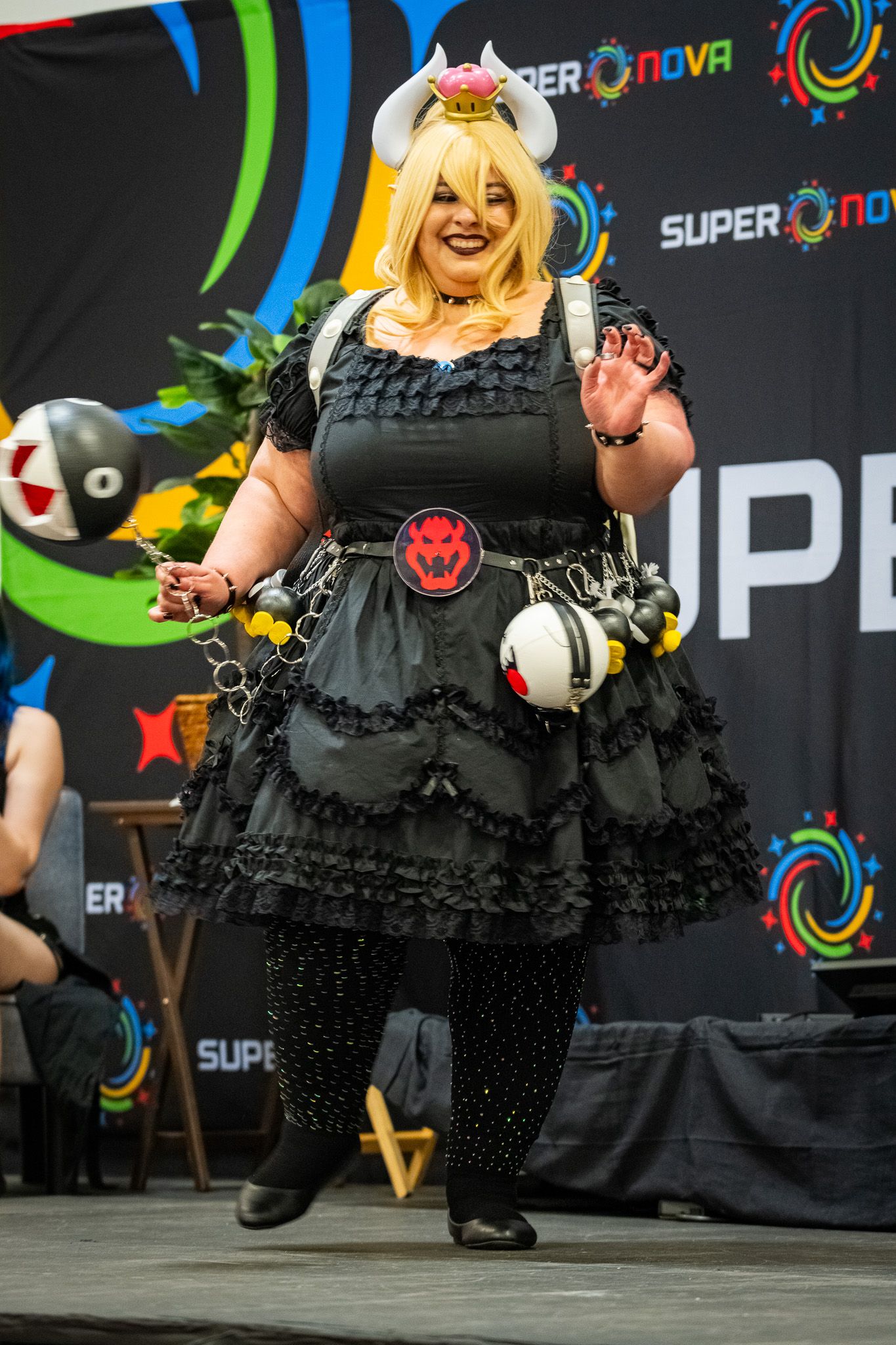 A cosplayer in a black gothic lolita dress poses in front of a gaming convention backdrop.