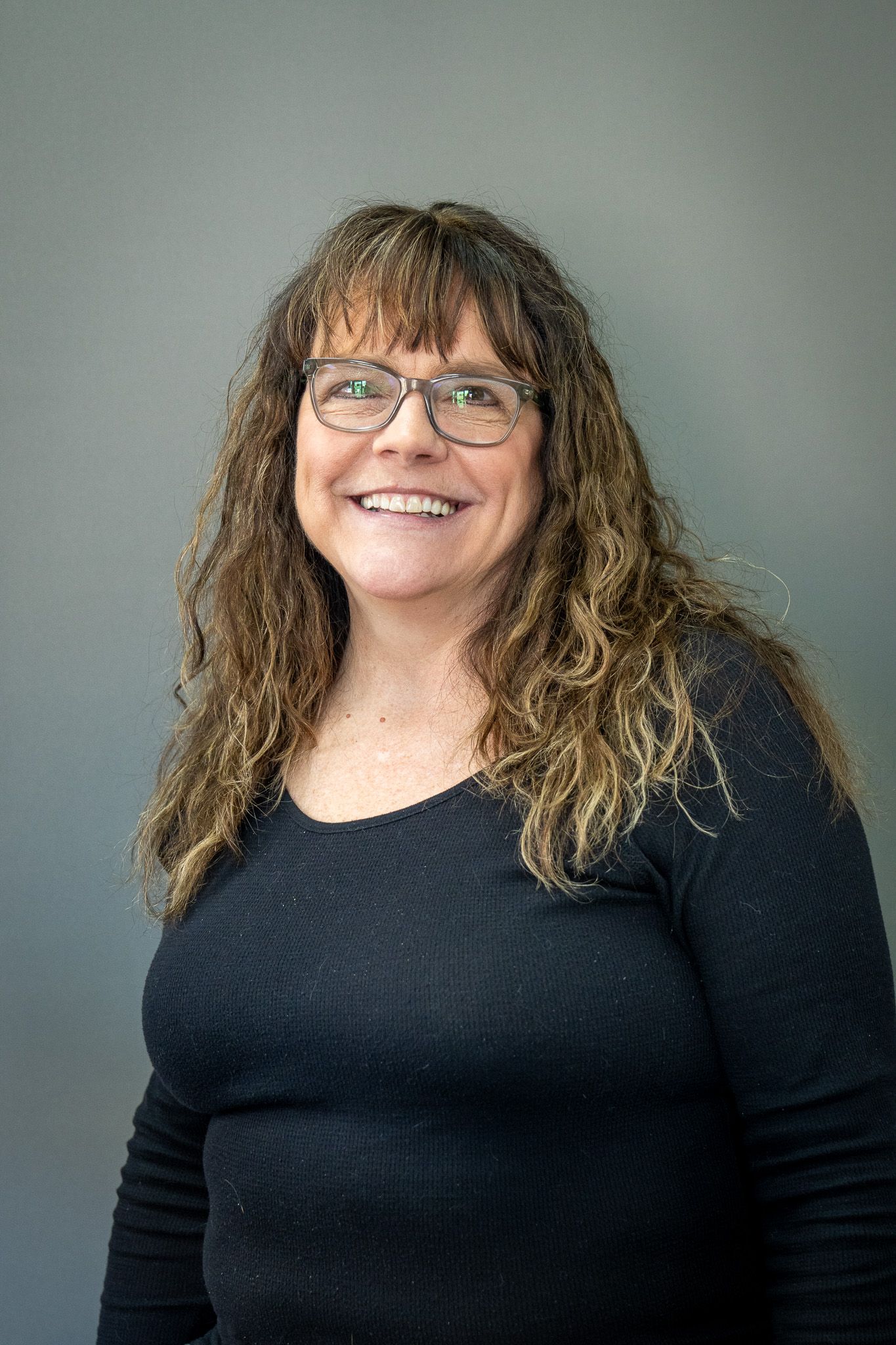 Professional headshot of person with curly brown hair, glasses and black sweater against grey background.