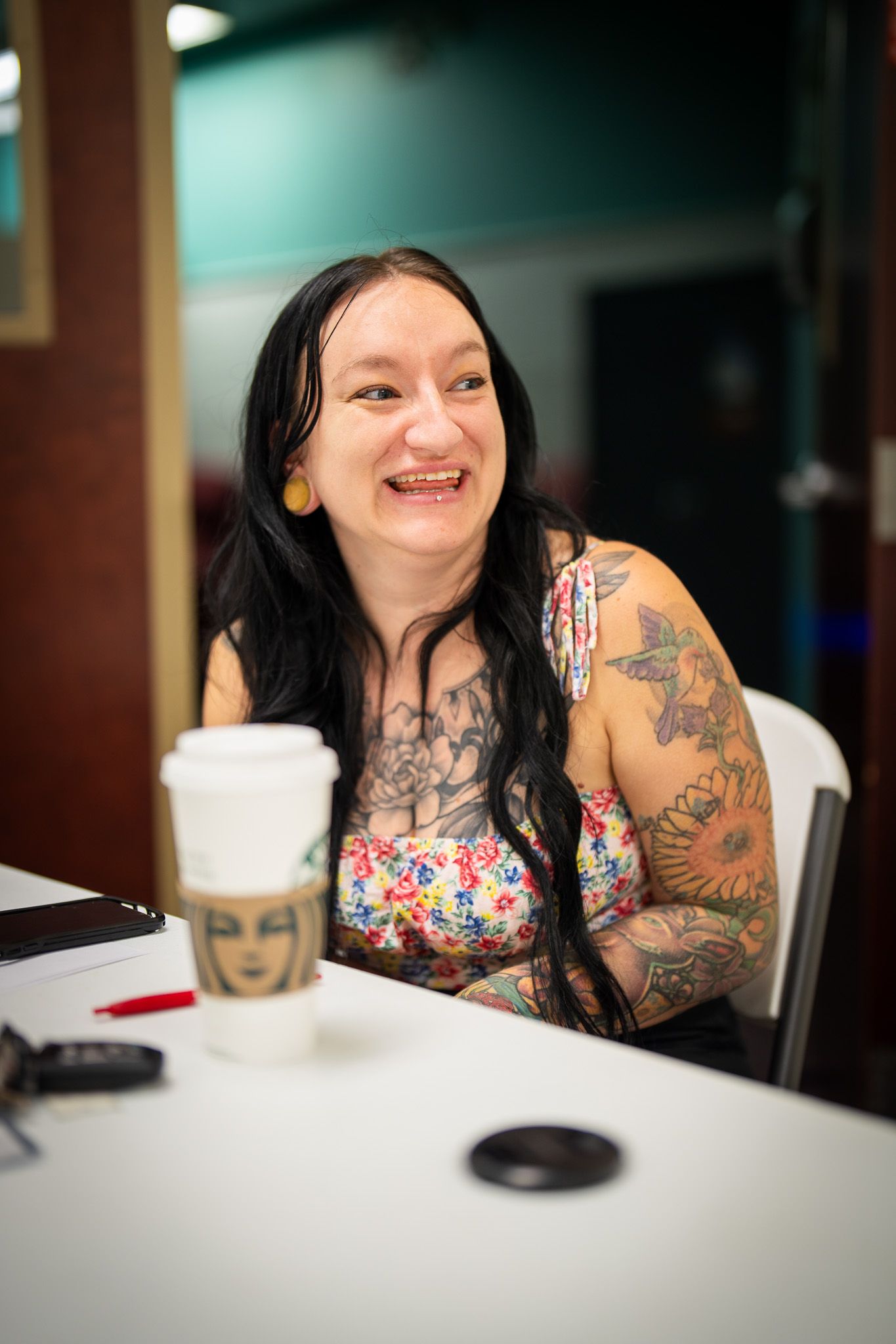 A tattooed person in floral top sits at white table with coffee cup while laughing joyfully.