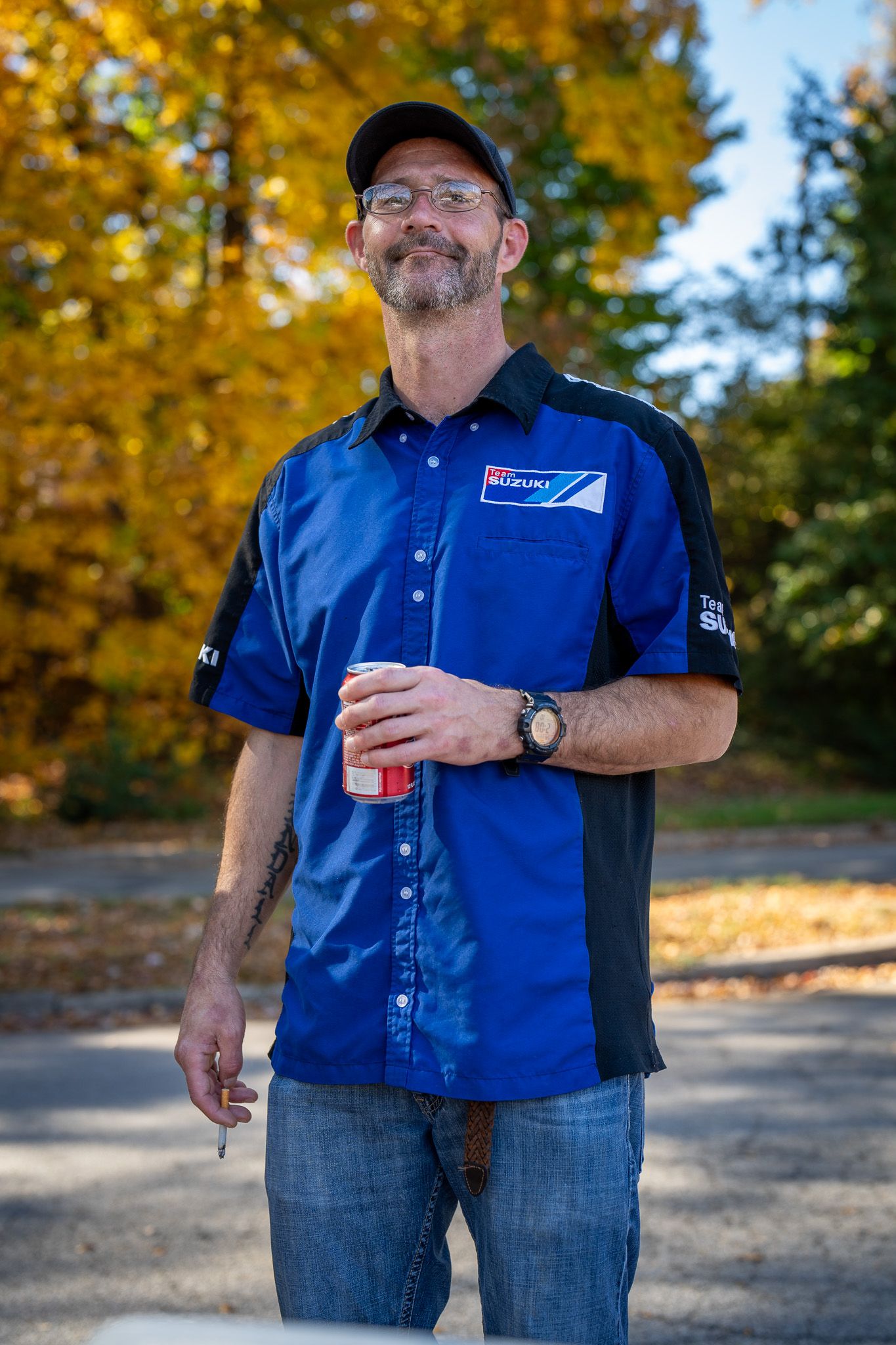 Employee in blue uniform shirt and baseball cap stands outdoors during autumn with fall foliage in background.