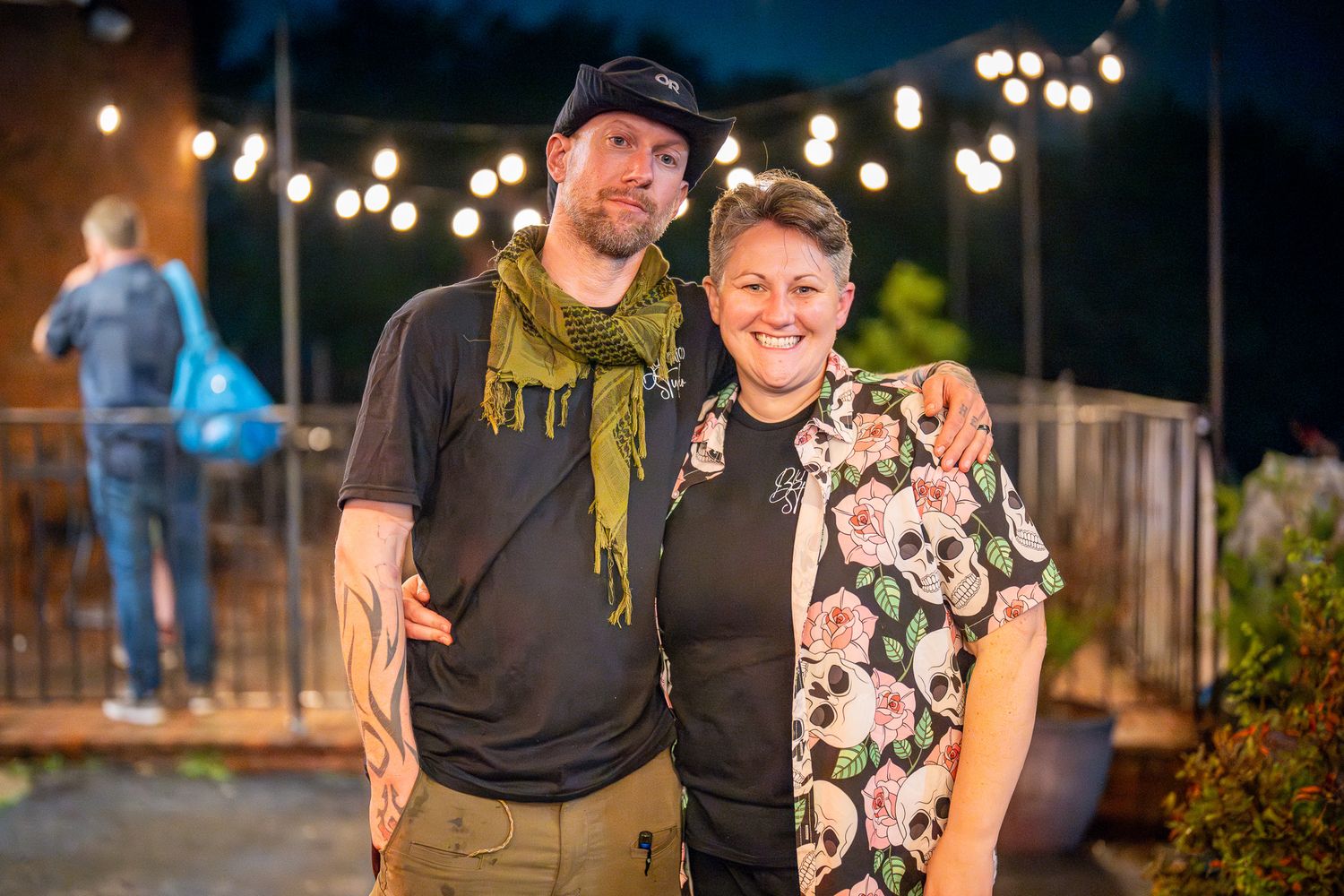 Two people stand together at an outdoor evening gathering with string lights glowing in the background.