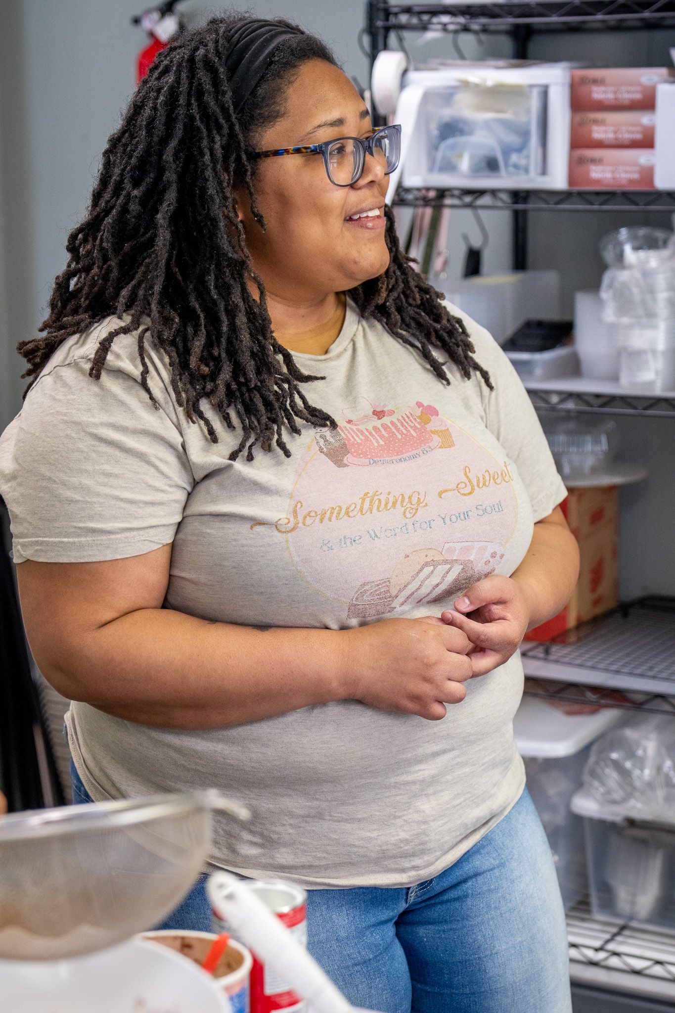 Small business owner wearing glasses and t-shirt stands in storeroom with metal shelving and storage containers.