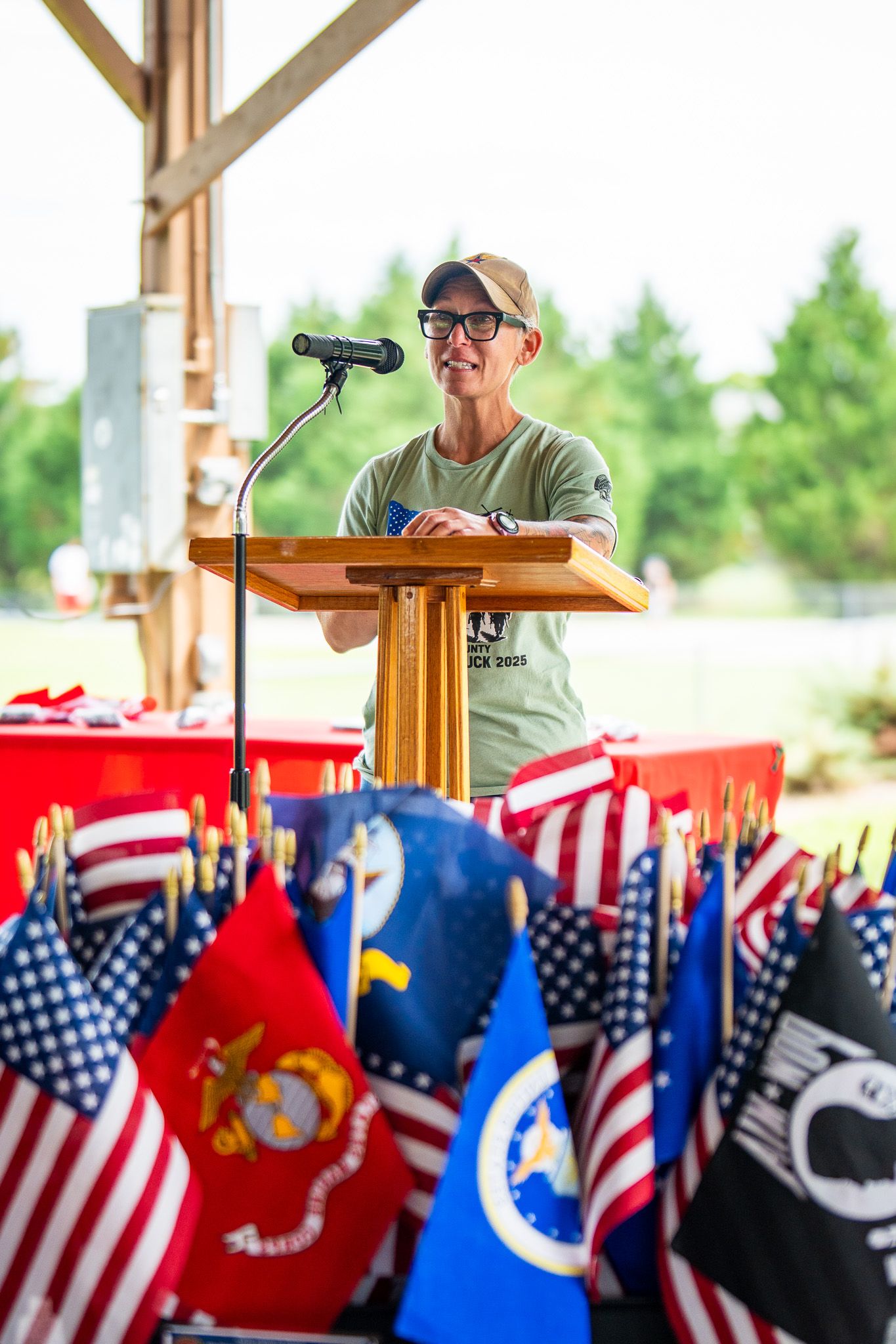 Speaker at wooden podium with American and military branch flags displayed in foreground at outdoor patriotic event.