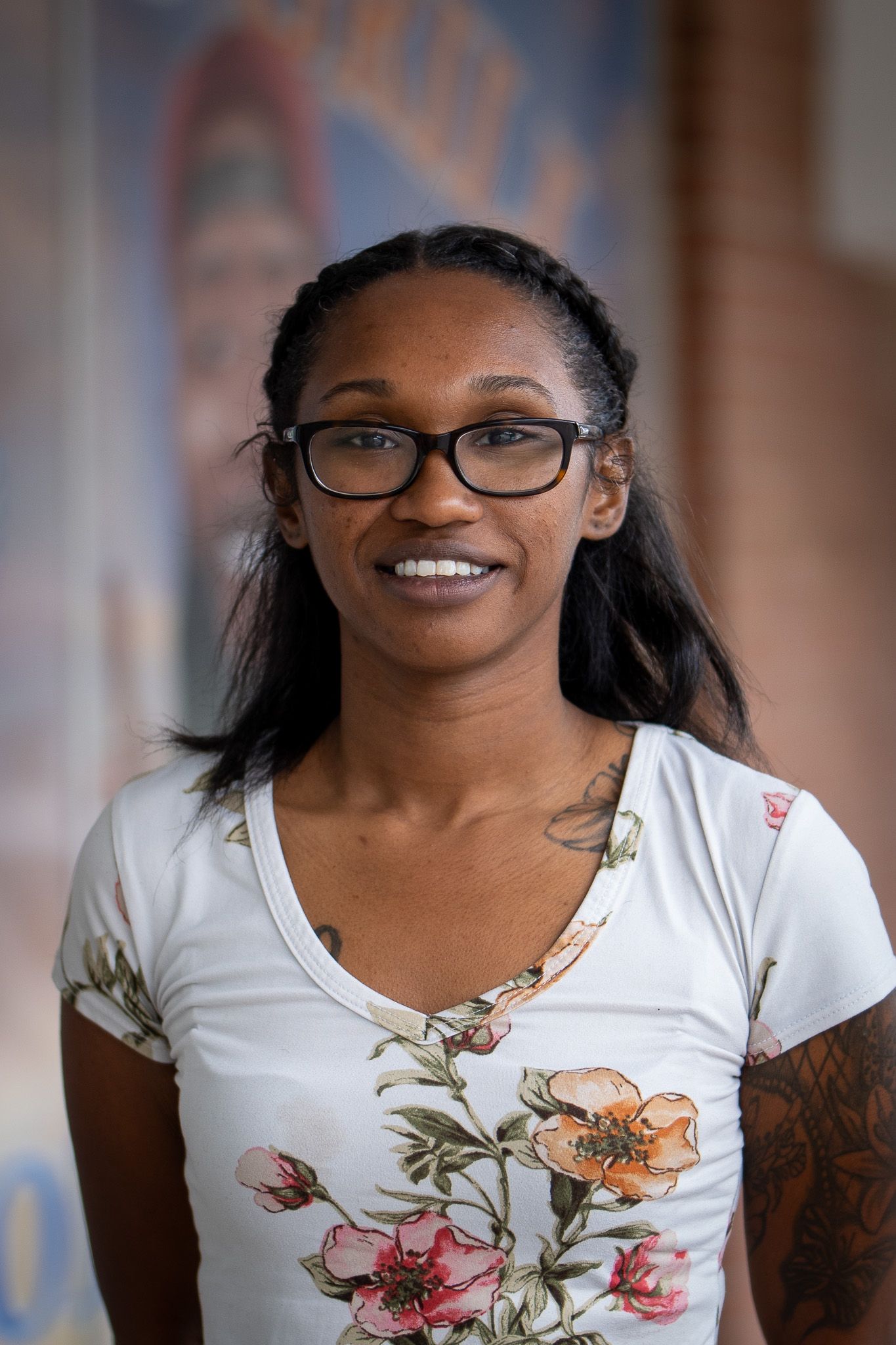 A person wearing glasses and a white floral print t-shirt smiles warmly at the camera against a blurred background.