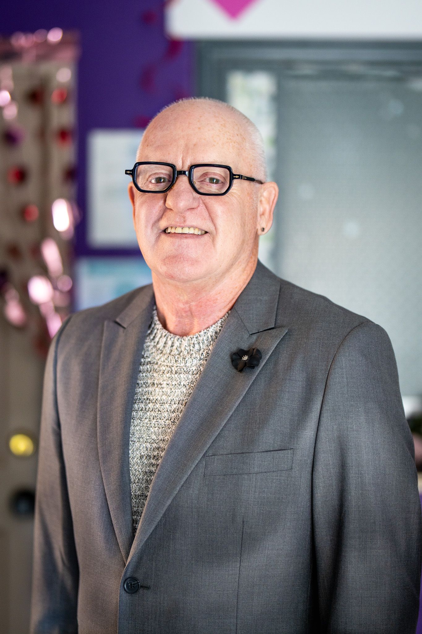 A person wearing glasses, a gray suit jacket and knit sweater stands smiling in an indoor setting with purple walls.