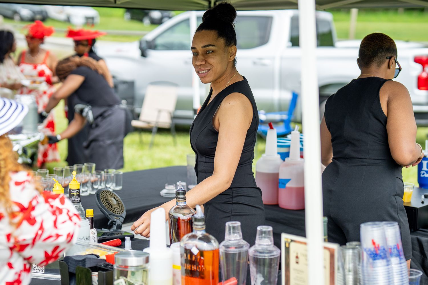 A bartender in a black tank top smiles while preparing drinks at an outdoor event with bottles displayed on the counter.