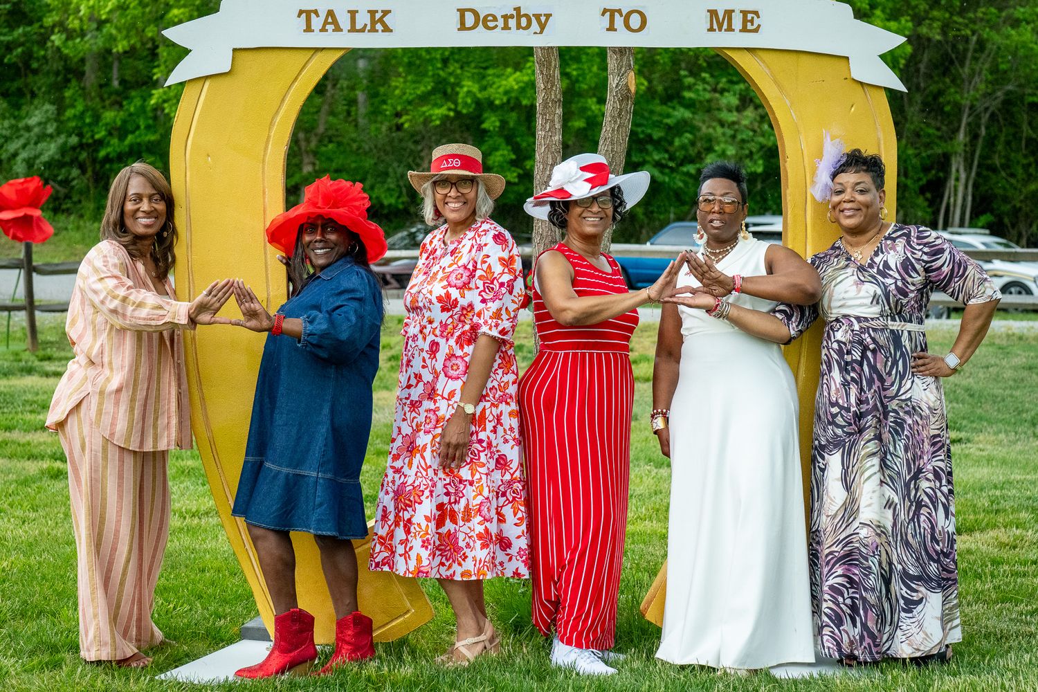A group of friends pose under a yellow 'Talk Derby To Me' arch wearing colorful dresses and hats at a garden party.