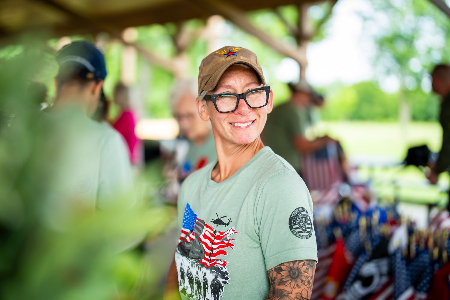 A person in a green t-shirt and baseball cap smiling at an outdoor event with tattoos visible on their arm.
