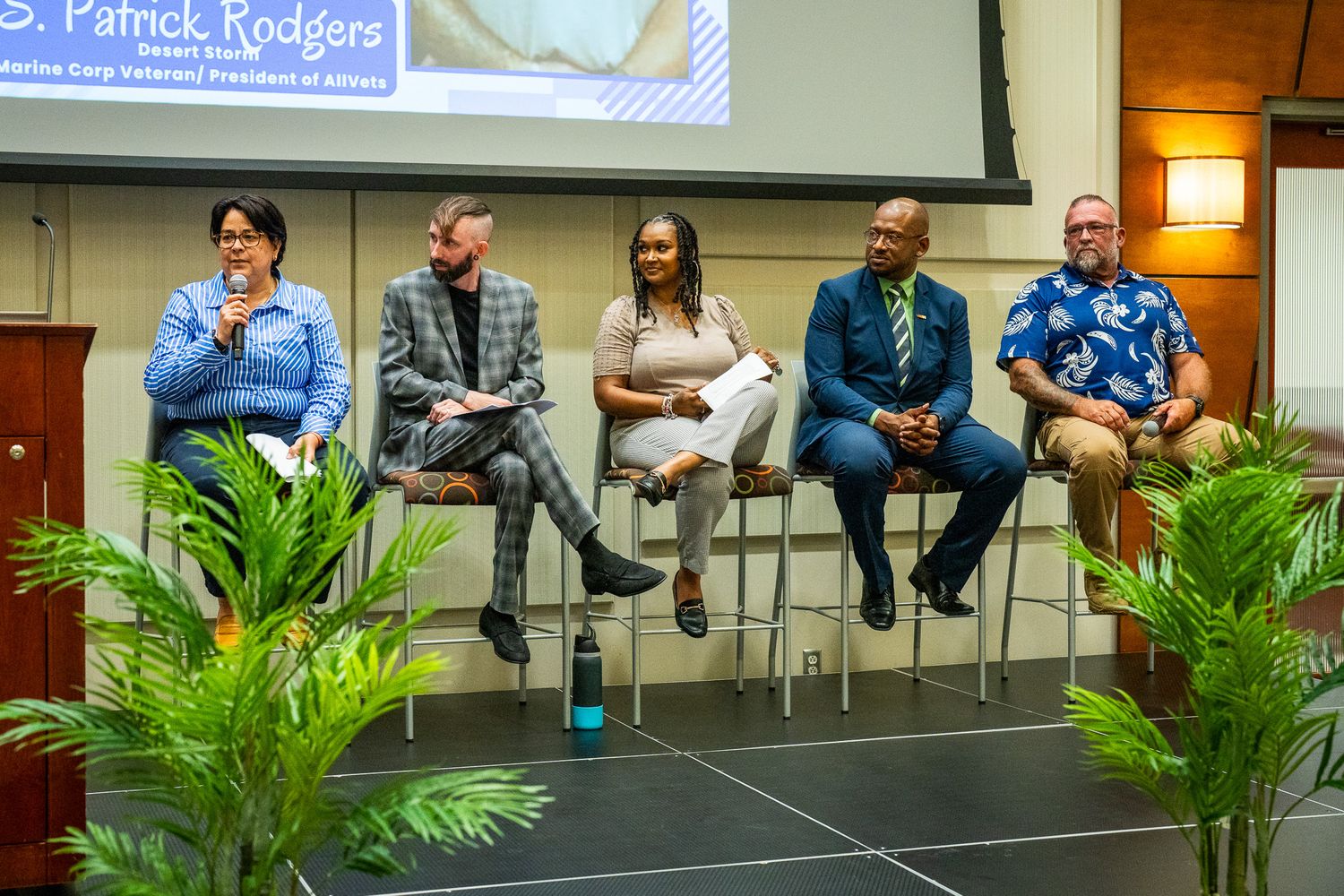 Panel discussion with five speakers seated on stage with potted plants and projector screen in background.
