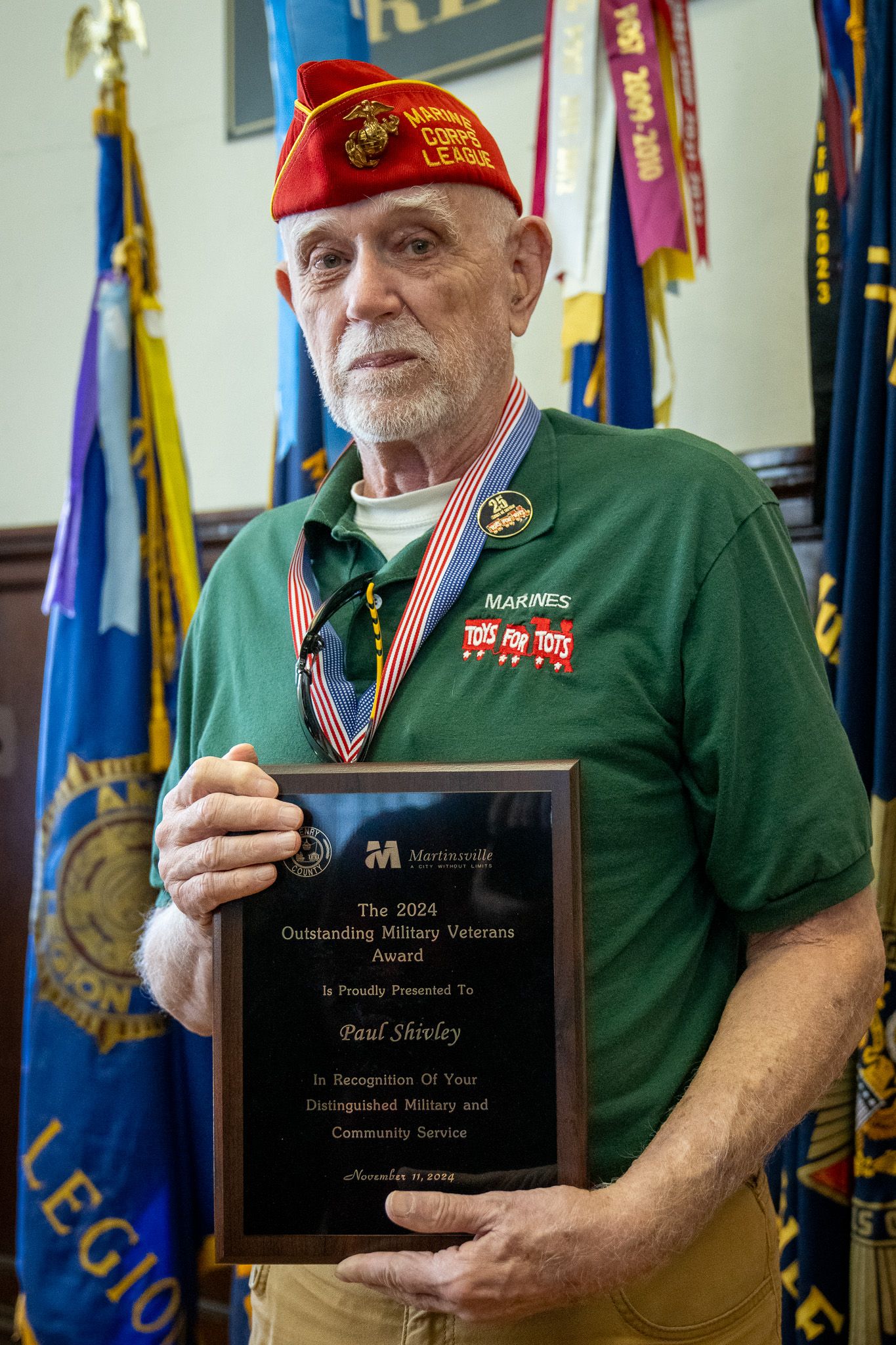 Military veterans display plaques and awards at a recognition ceremony with flags and banners in the background.