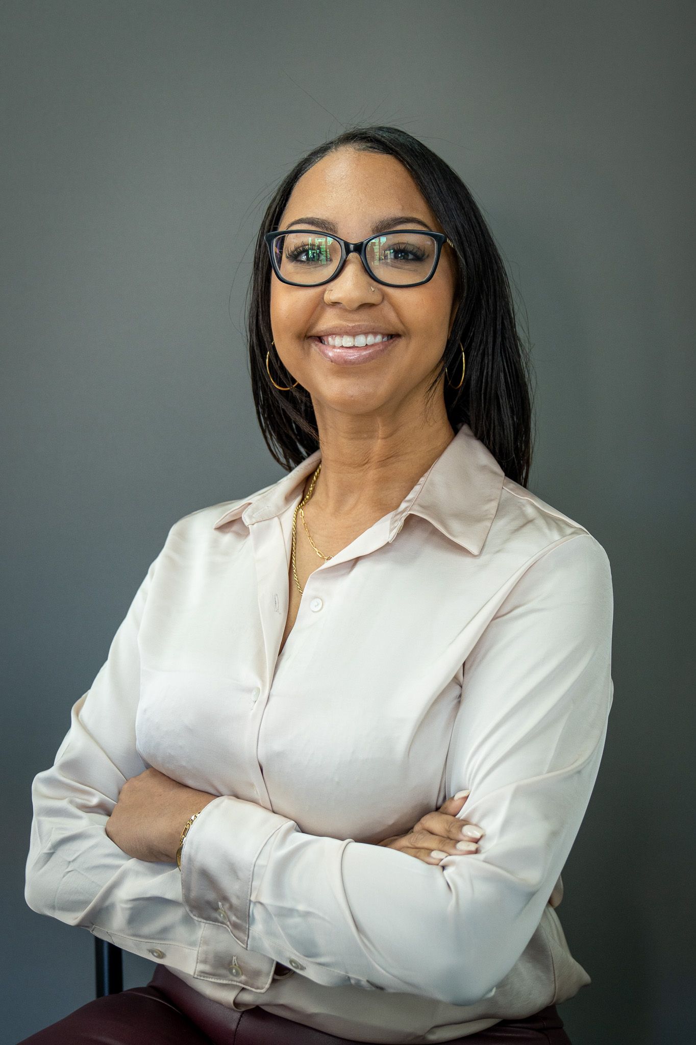 Professional headshot of a businesswoman in glasses and white shirt smiling with arms crossed against gray background.