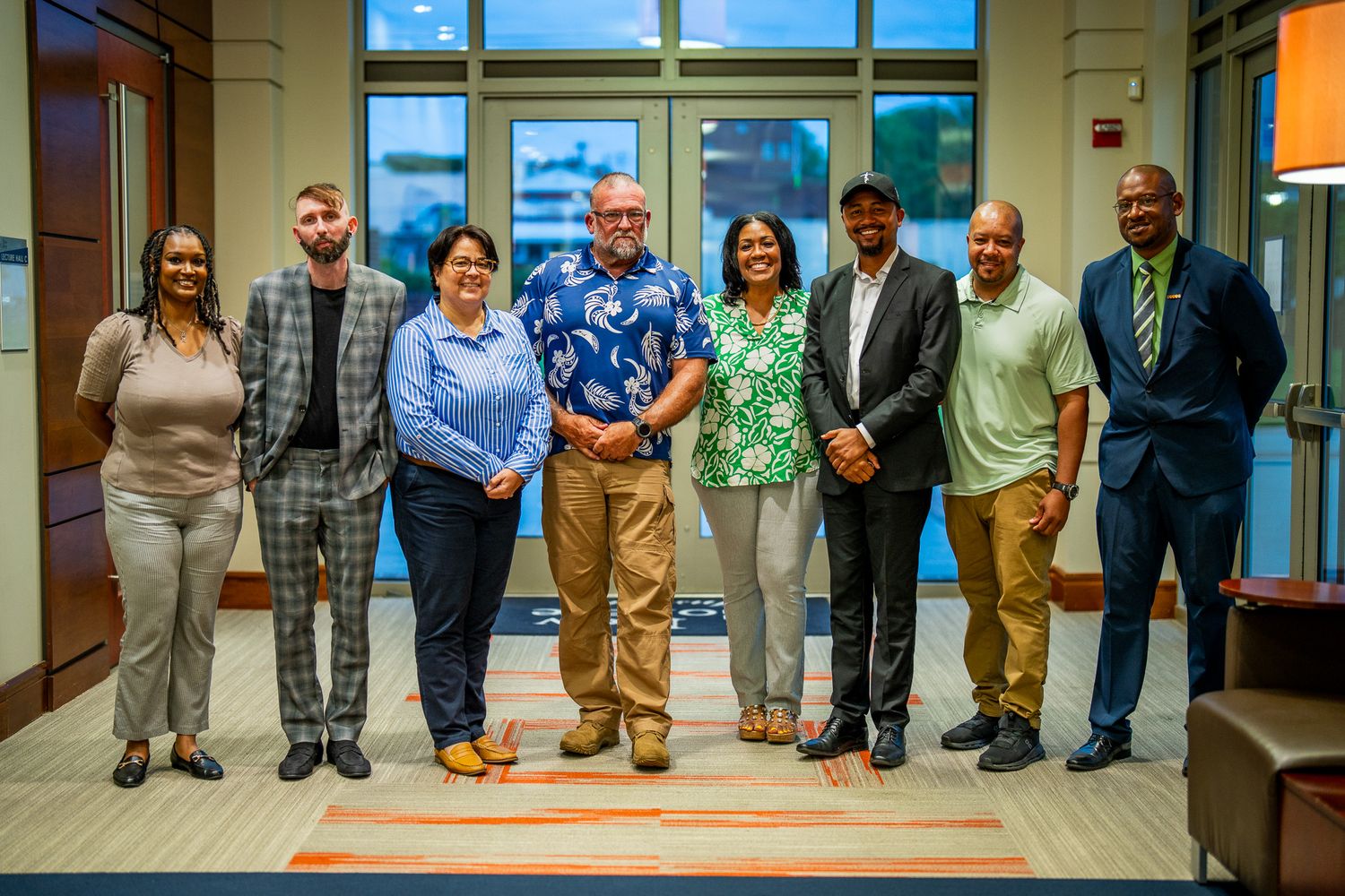 Eight professionals pose for a group photo in front of large windows in a contemporary office setting.