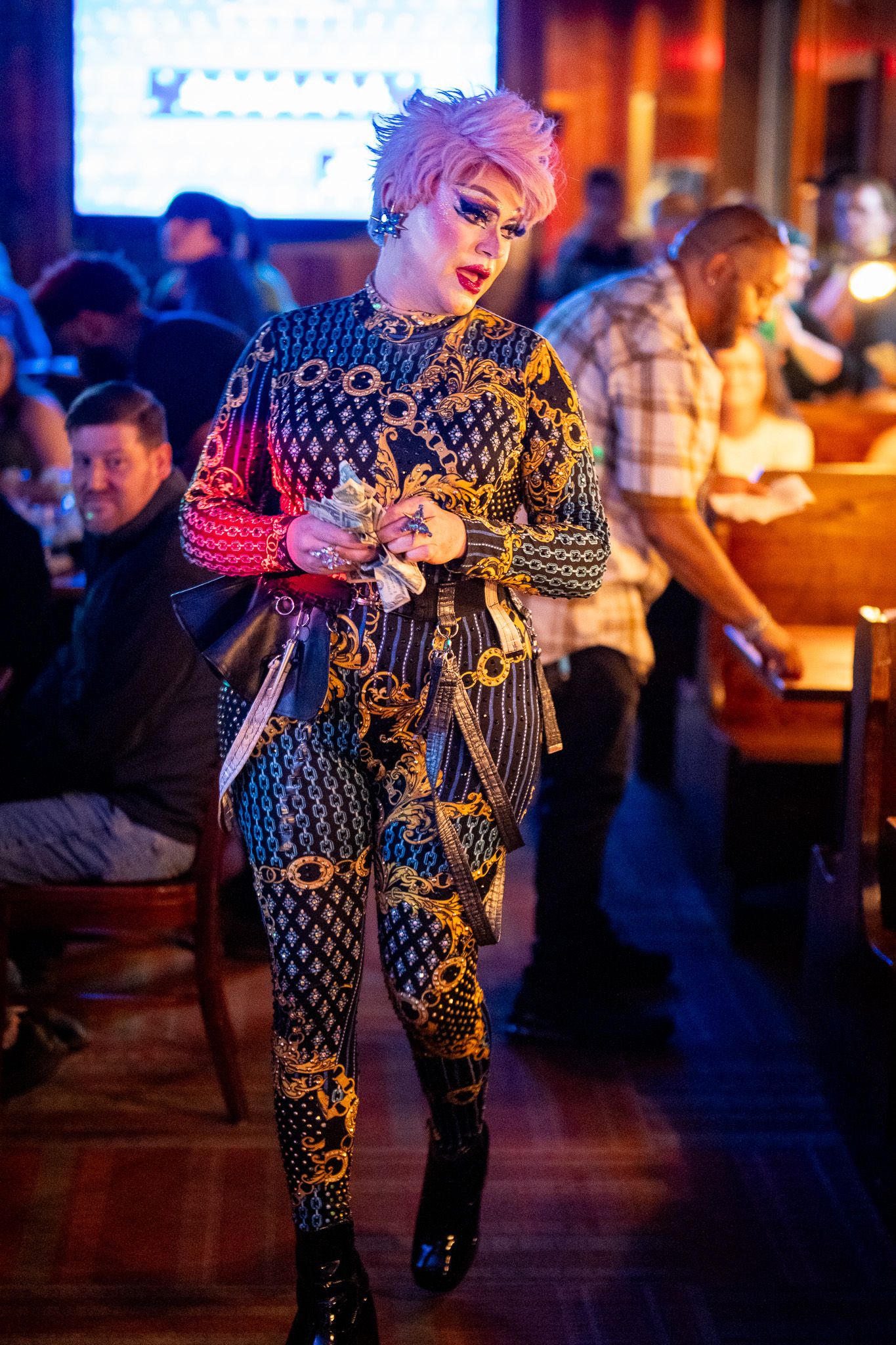 Performer wearing sparkly black and gold patterned jumpsuit strikes pose in dimly lit bar setting.