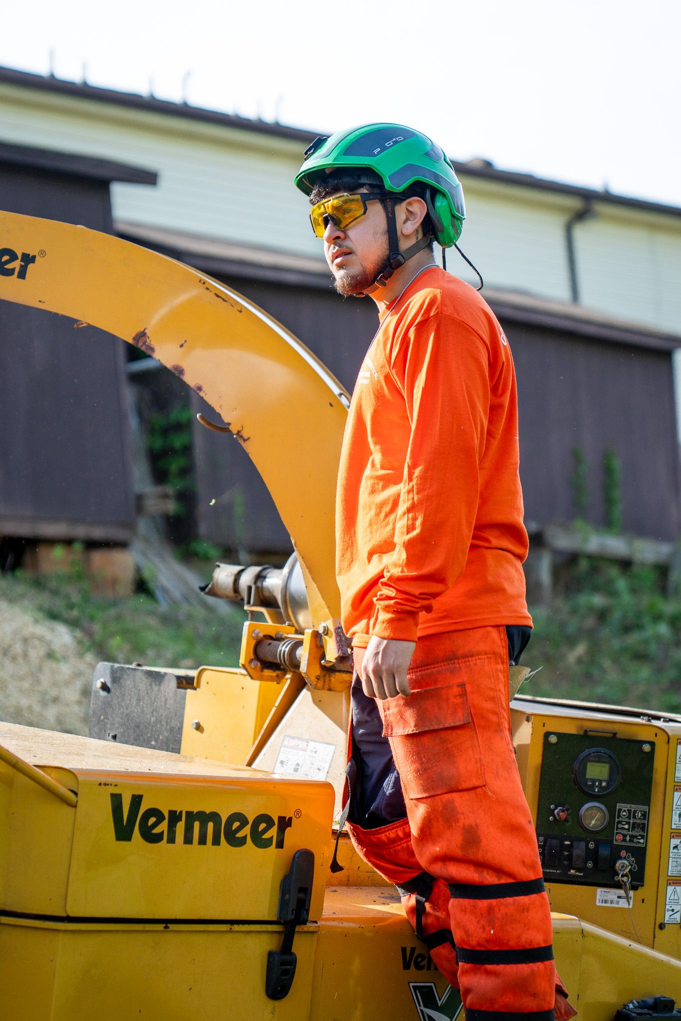 A worker in bright orange safety gear and green helmet operates a yellow Vermeer wood chipper machine.