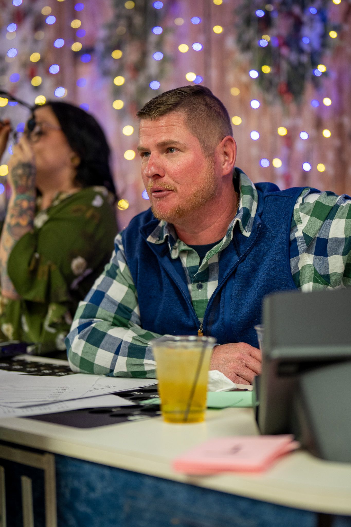 A person in a blue vest and plaid shirt sits at a desk with twinkle lights in the background during an event.
