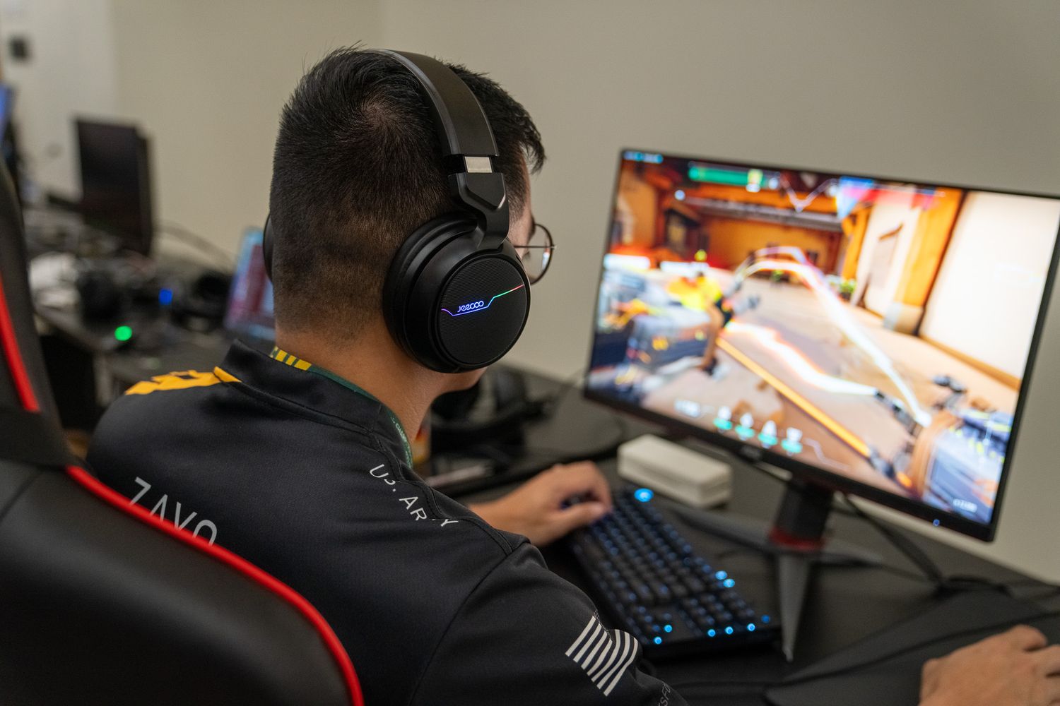 Gamer wearing black headphones sits in gaming chair while playing competitive video game on large monitor.