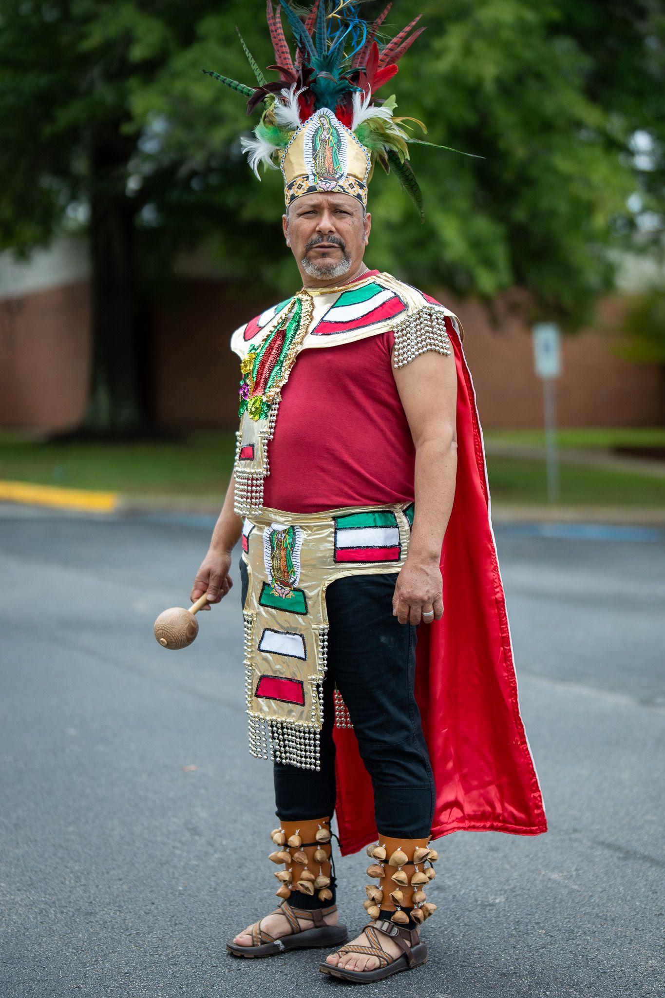 Person wearing traditional Aztec-inspired ceremonial costume with feathered headdress, red cape and decorative regalia in parking lot.