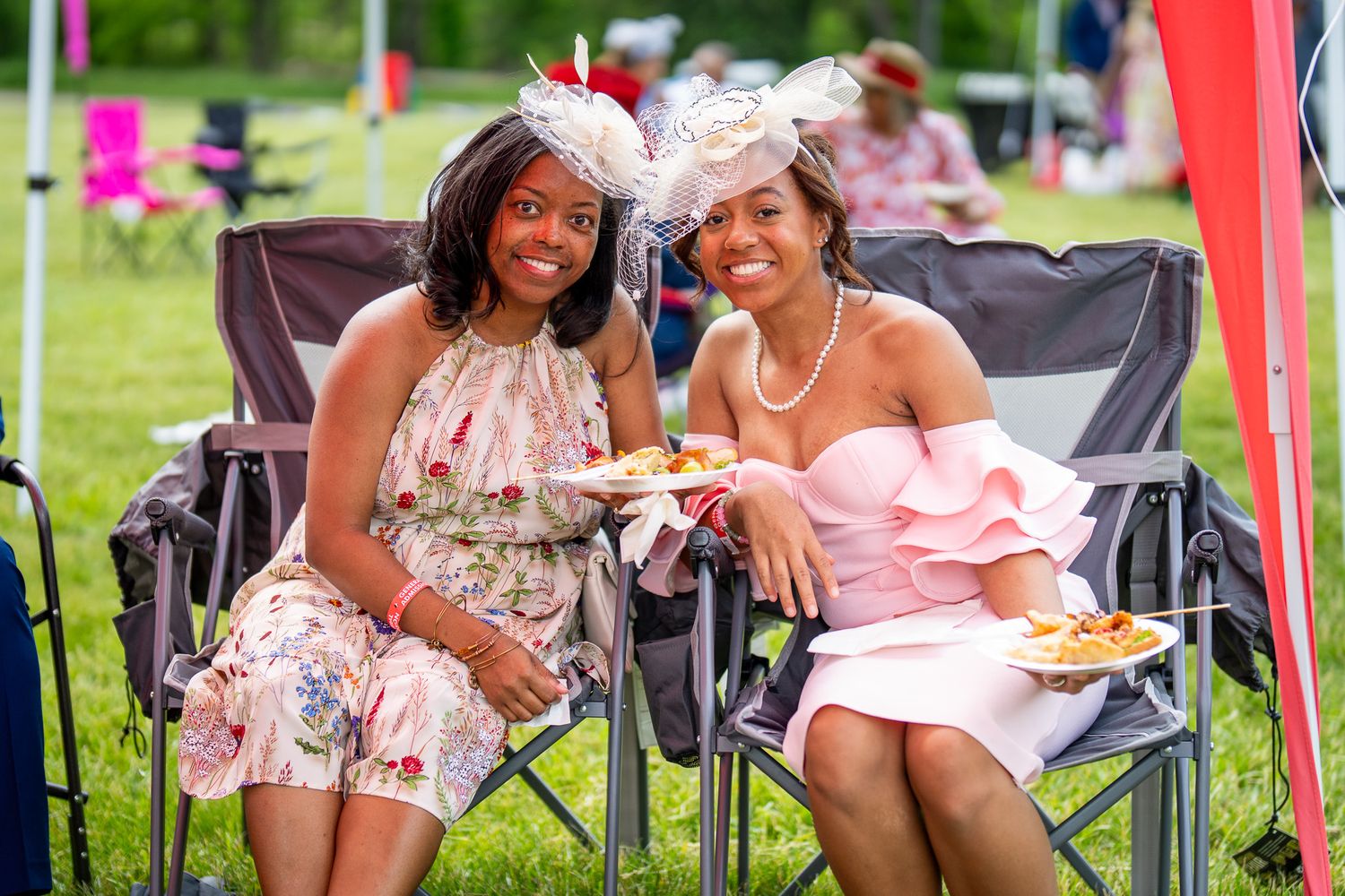 Two friends dressed in floral dresses and fancy hats enjoy refreshments together at an outdoor summer event.