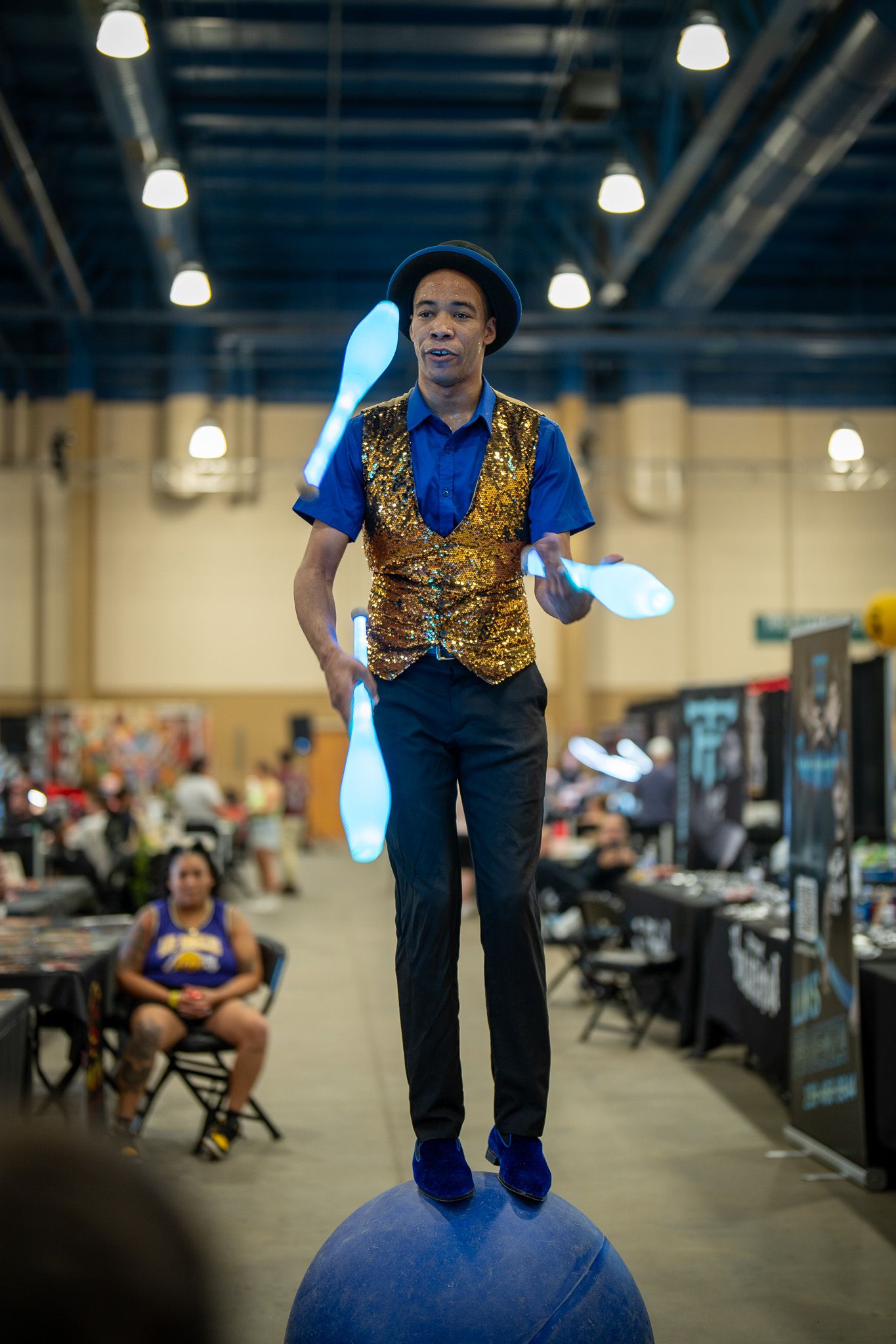 A performer in a gold sequined vest balances skillfully atop a blue ball while juggling in an indoor event space.