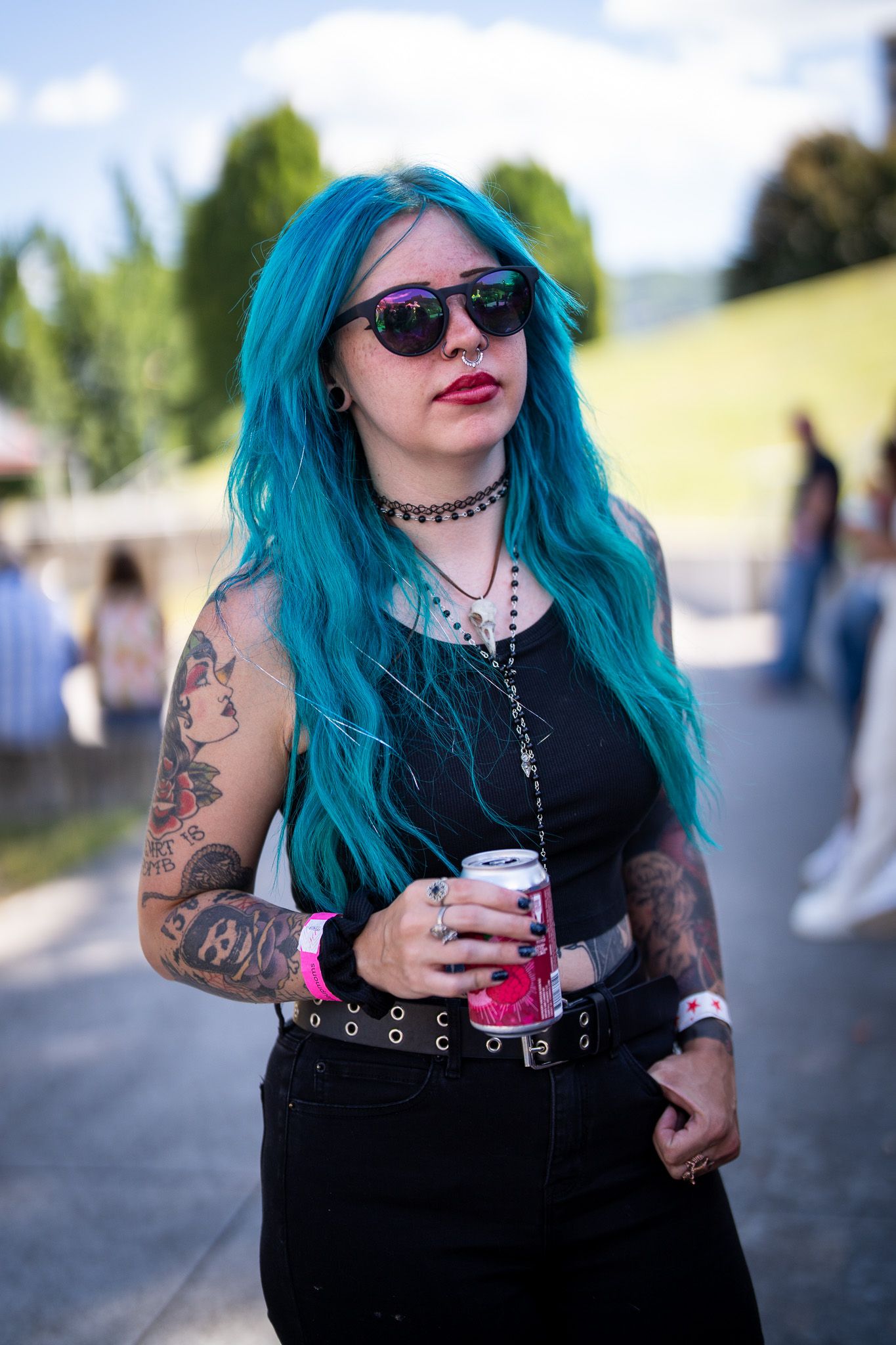 Alternative style with bright turquoise hair, black clothing, sunglasses and tattoos holds beverage at outdoor event.