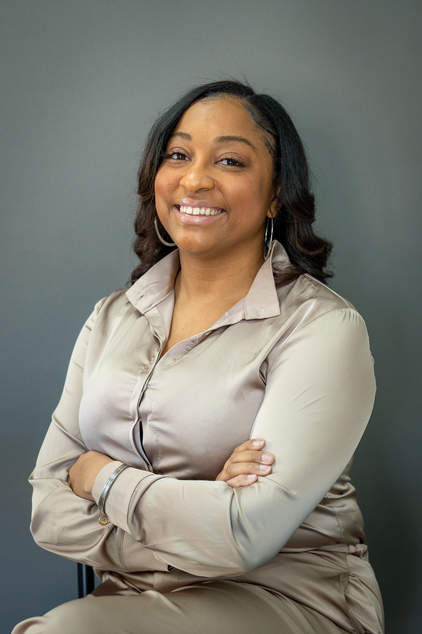 Corporate portrait of a businesswoman in beige blouse smiling warmly with crossed arms against neutral backdrop.