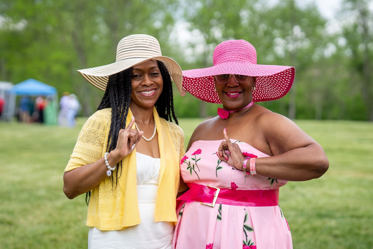 Two women wearing wide-brimmed sun hats enjoy an outdoor event in spring, one in yellow and one in pink attire.
