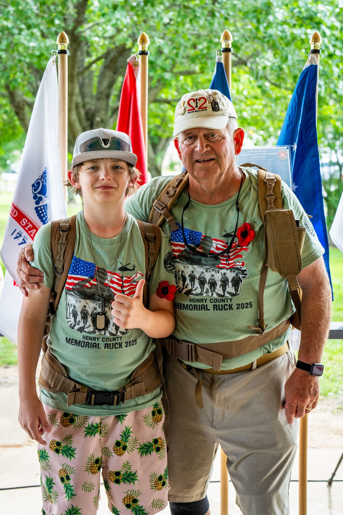 Two people wearing matching military-inspired t-shirts pose together in front of American flags during a memorial event.