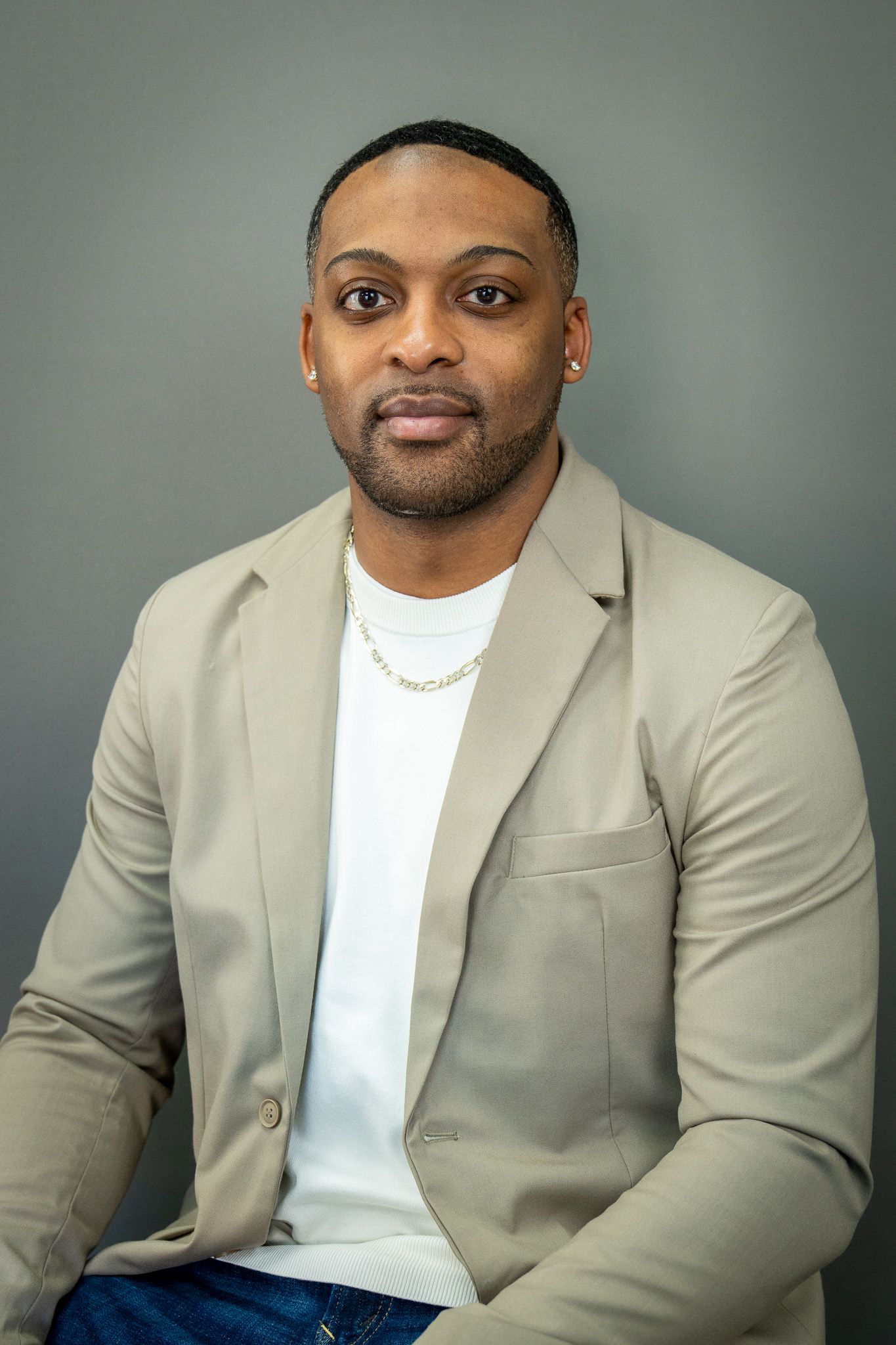 Professional headshot of person wearing a beige blazer with white t-shirt against grey background.