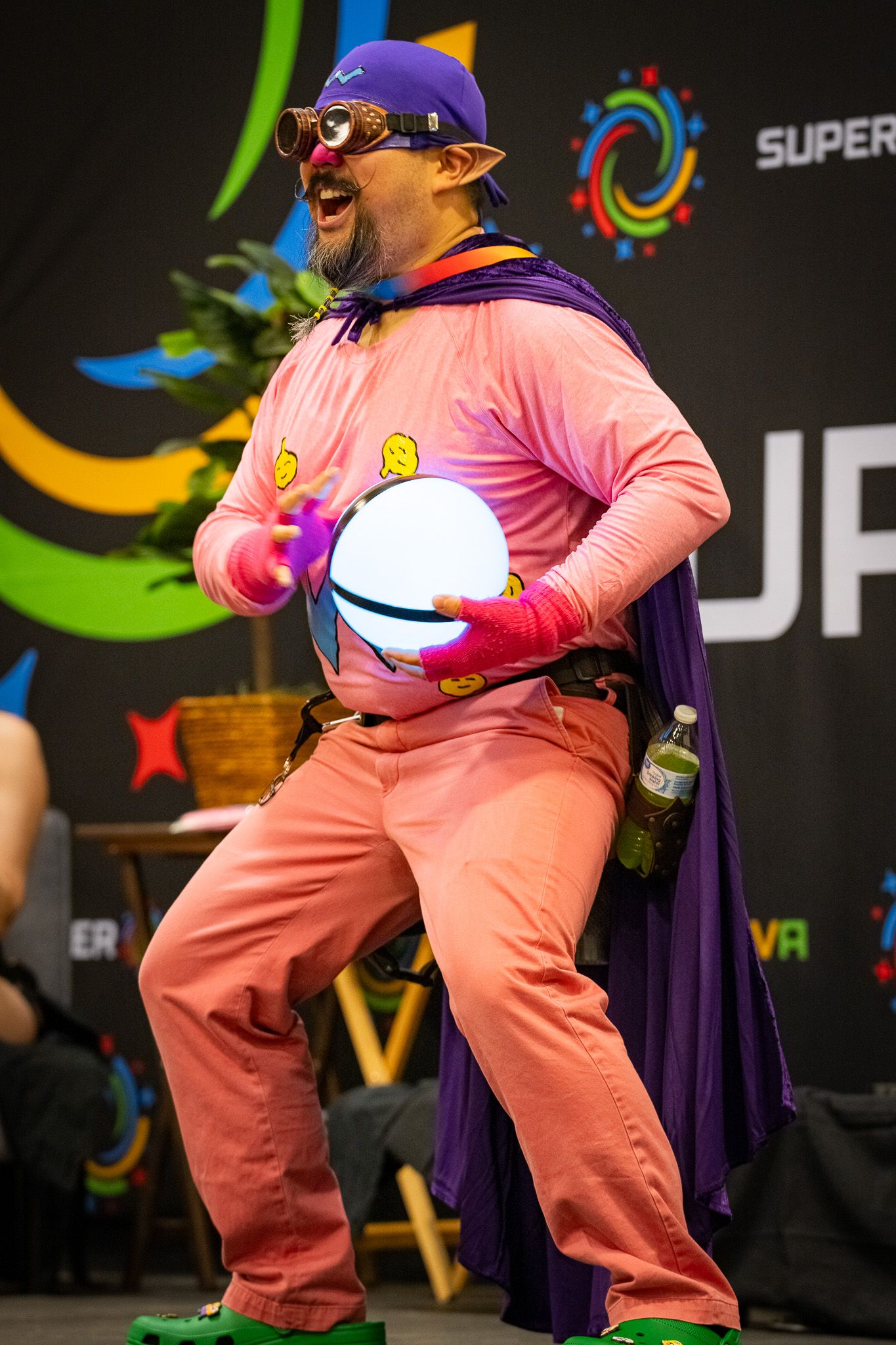 A superhero cosplayer in pink outfit and purple cape poses energetically on stage.