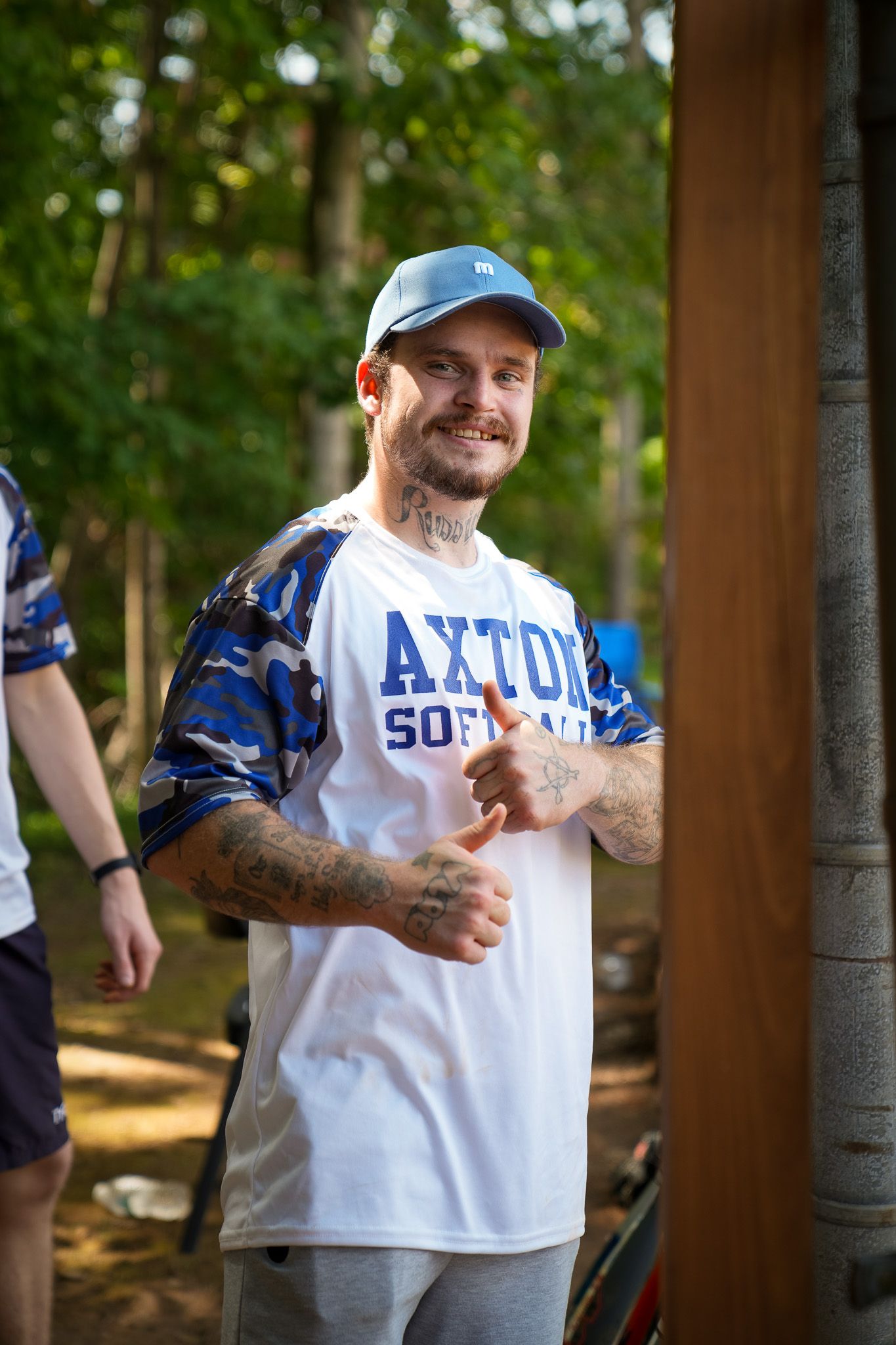Athletic person in a white t-shirt and baseball cap gives thumbs up while standing outdoors, showing tattoo-covered arms.