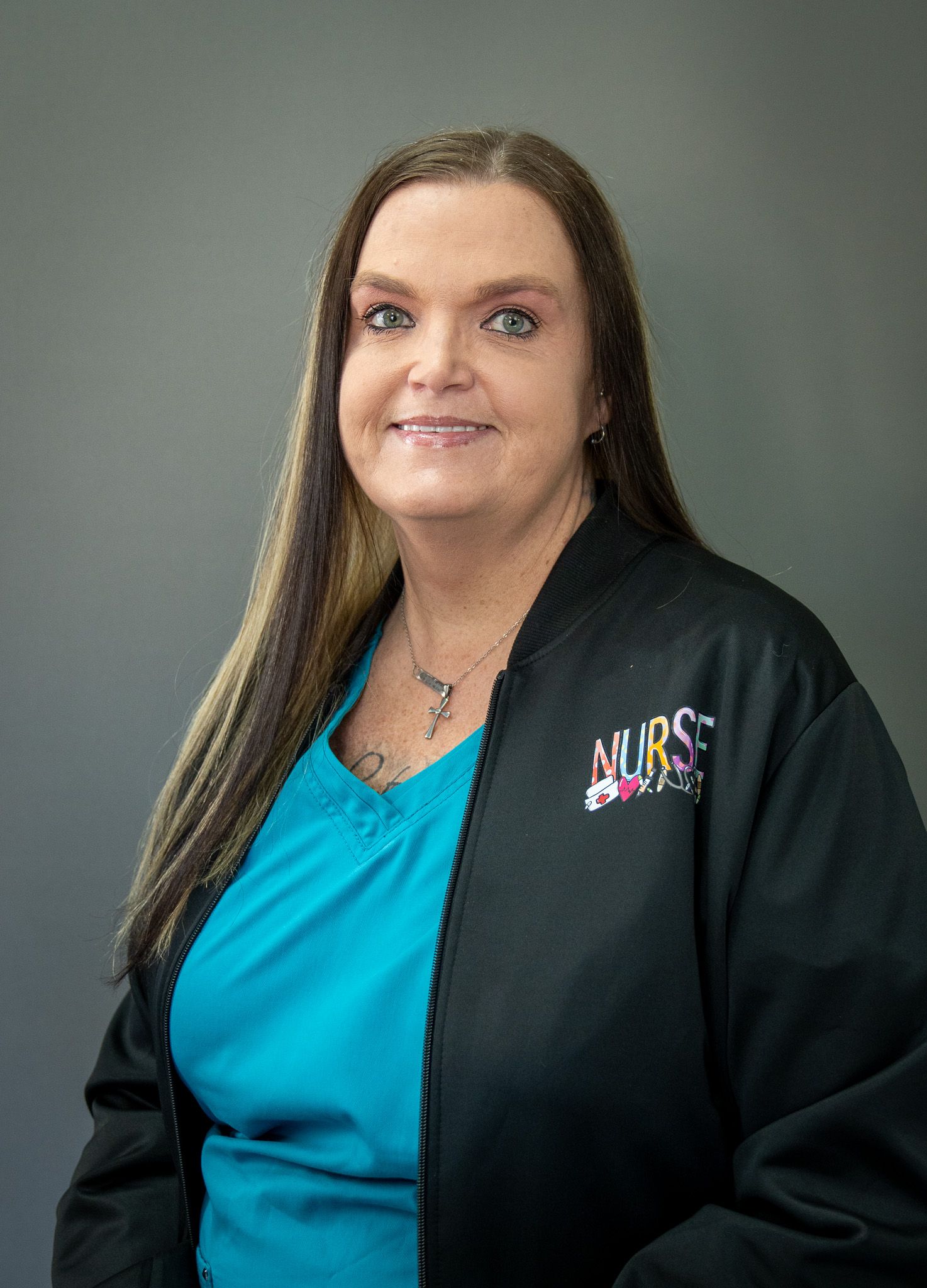 Medical professional in teal scrubs and black jacket with Nurse logo smiling against gray background.