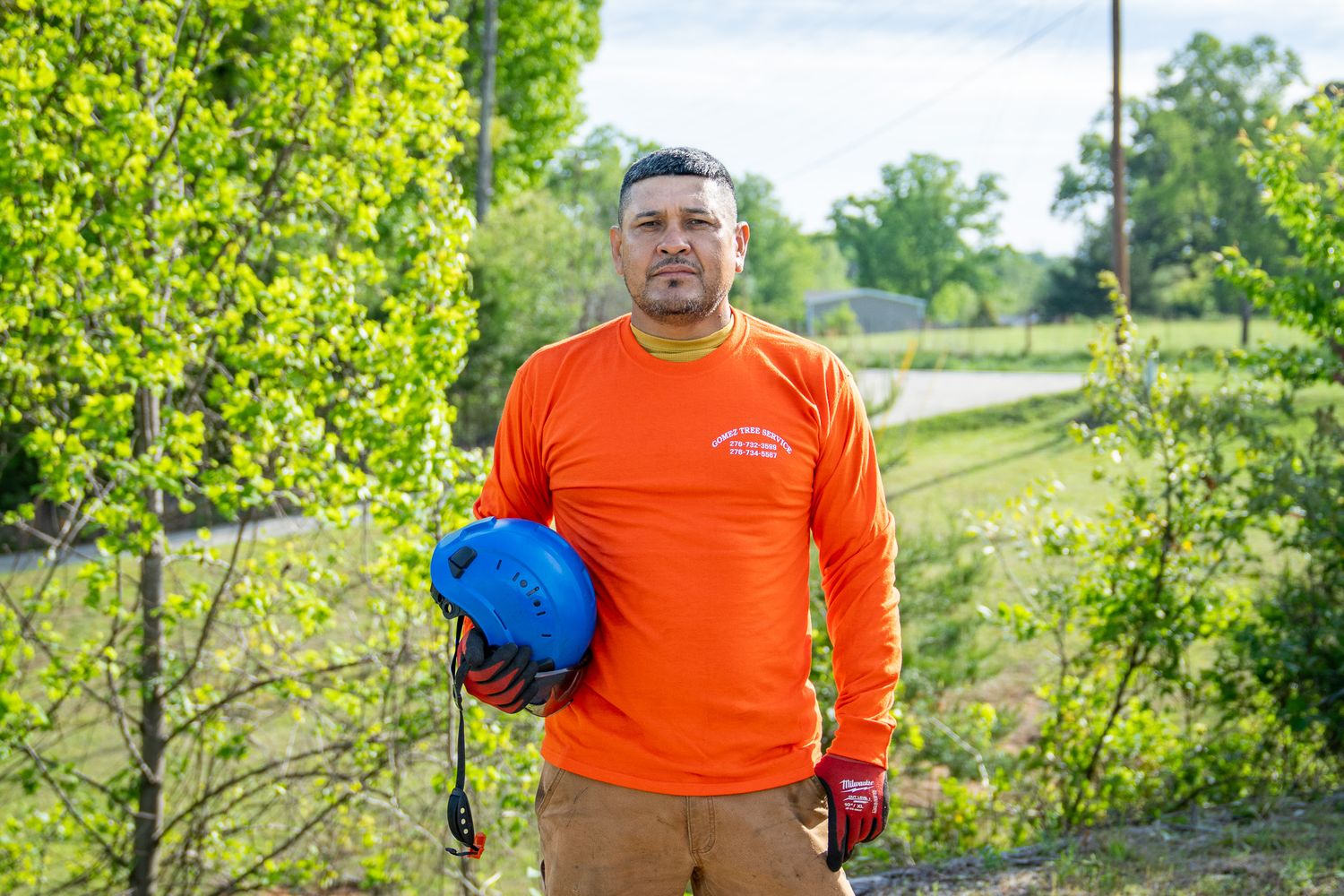Construction worker in bright orange safety shirt holding blue hard hat stands on outdoor path near green foliage.