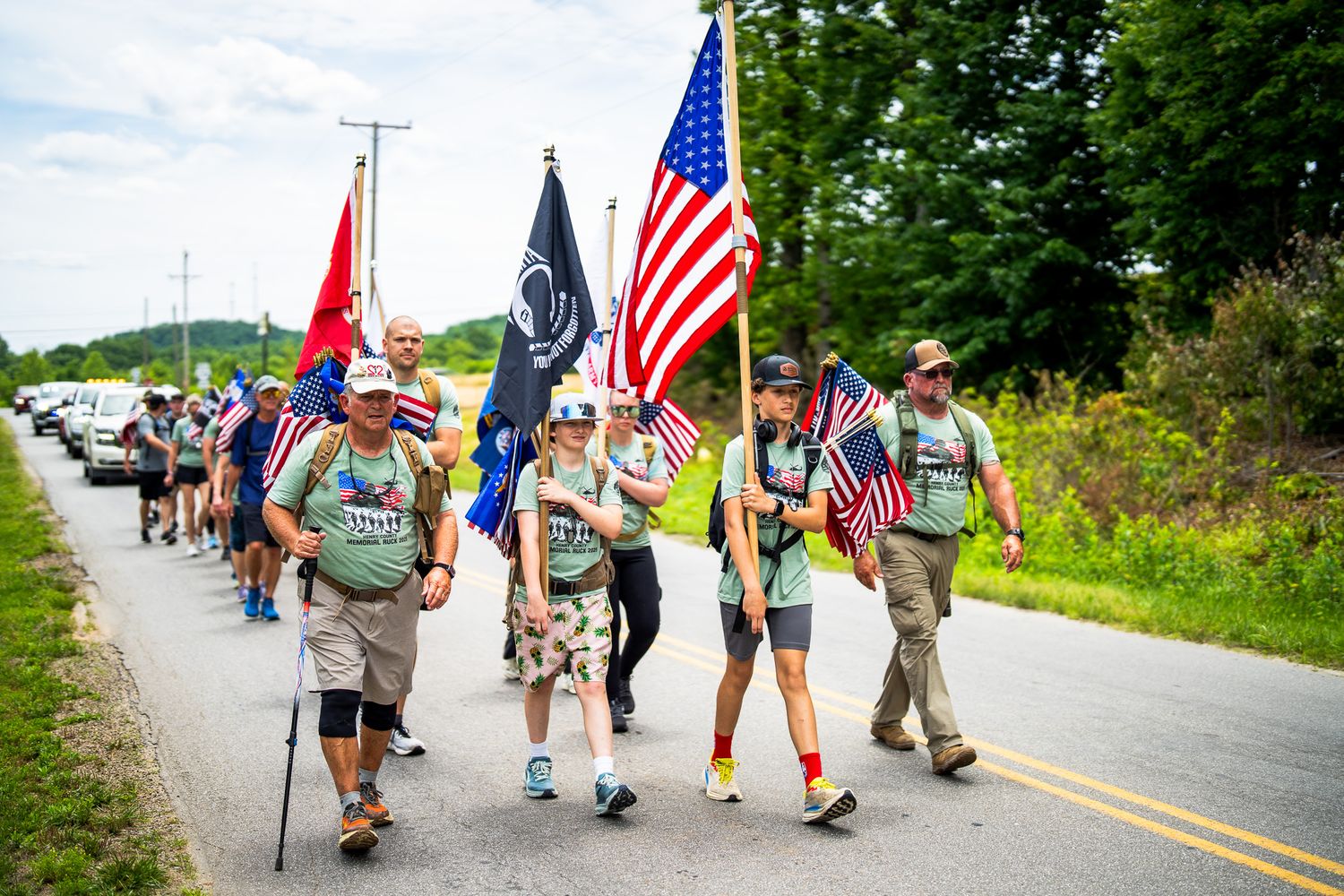 People walk together along a rural road carrying flags including the American flag on a sunny day.