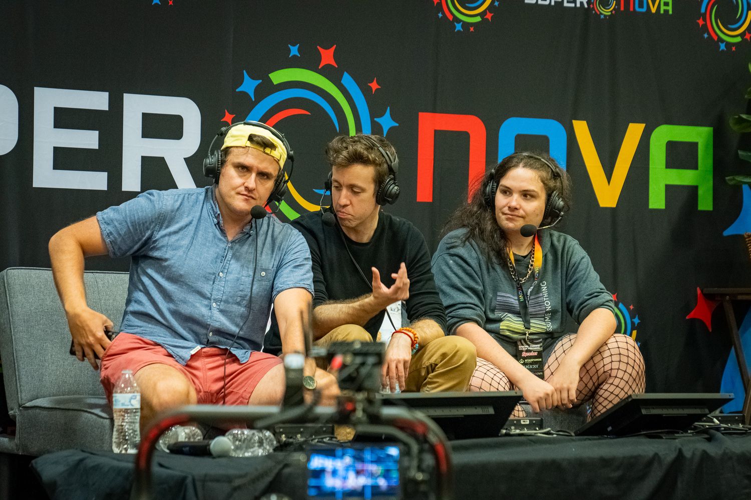 Three people sit on a stage with rainbow-colored graphics and text behind them at a gaming event.